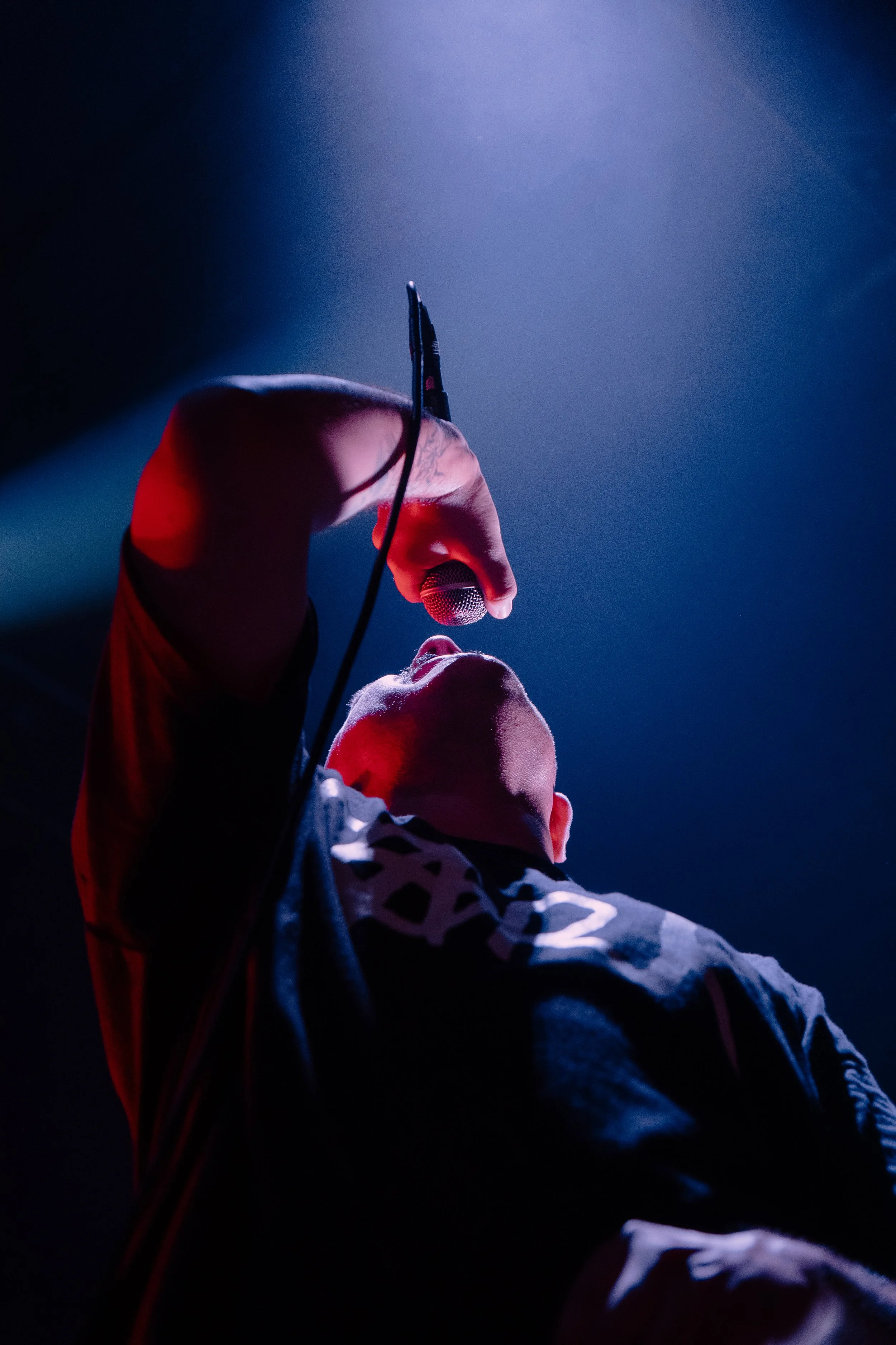 A performer on stage holding a microphone, with dramatic lighting and dressed in a patterned jacket, looking upward.