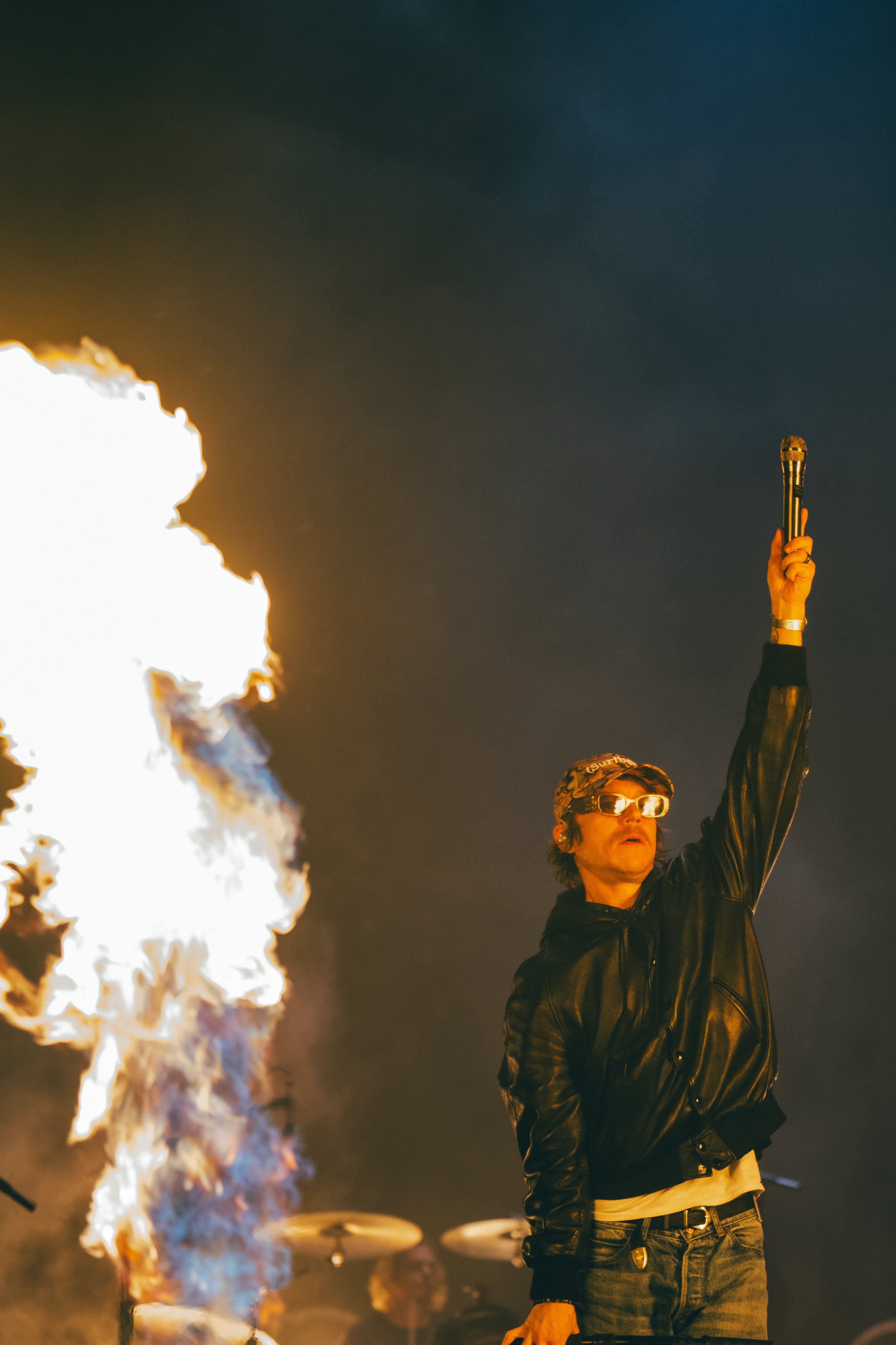 A person wearing glasses and a leather jacket holding a microphone with a fire burning in the background at night.