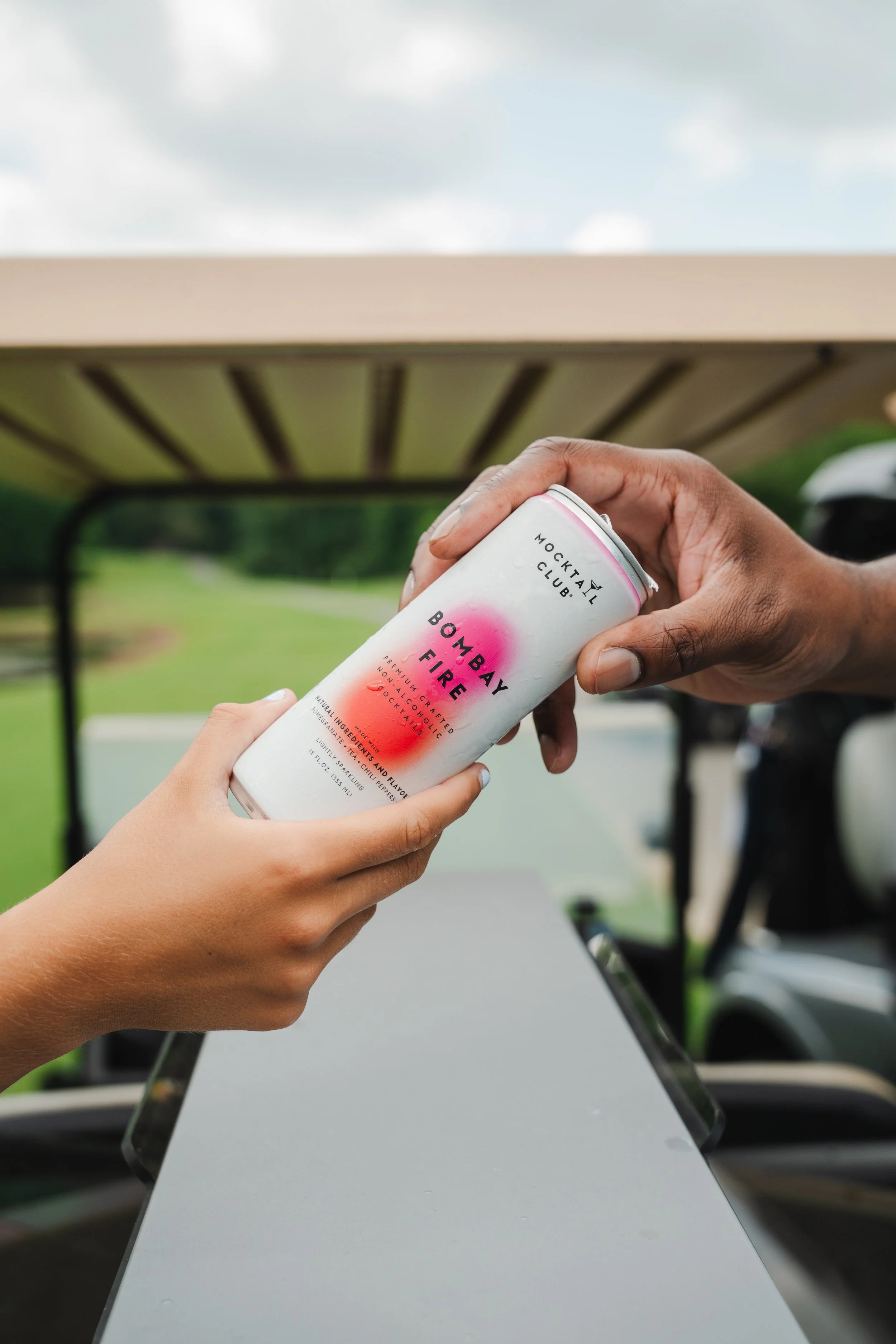 A person holding a can of Bombay Fire Mango flavored canned cocktail with a blurred golf cart and outdoor scenery in the background.