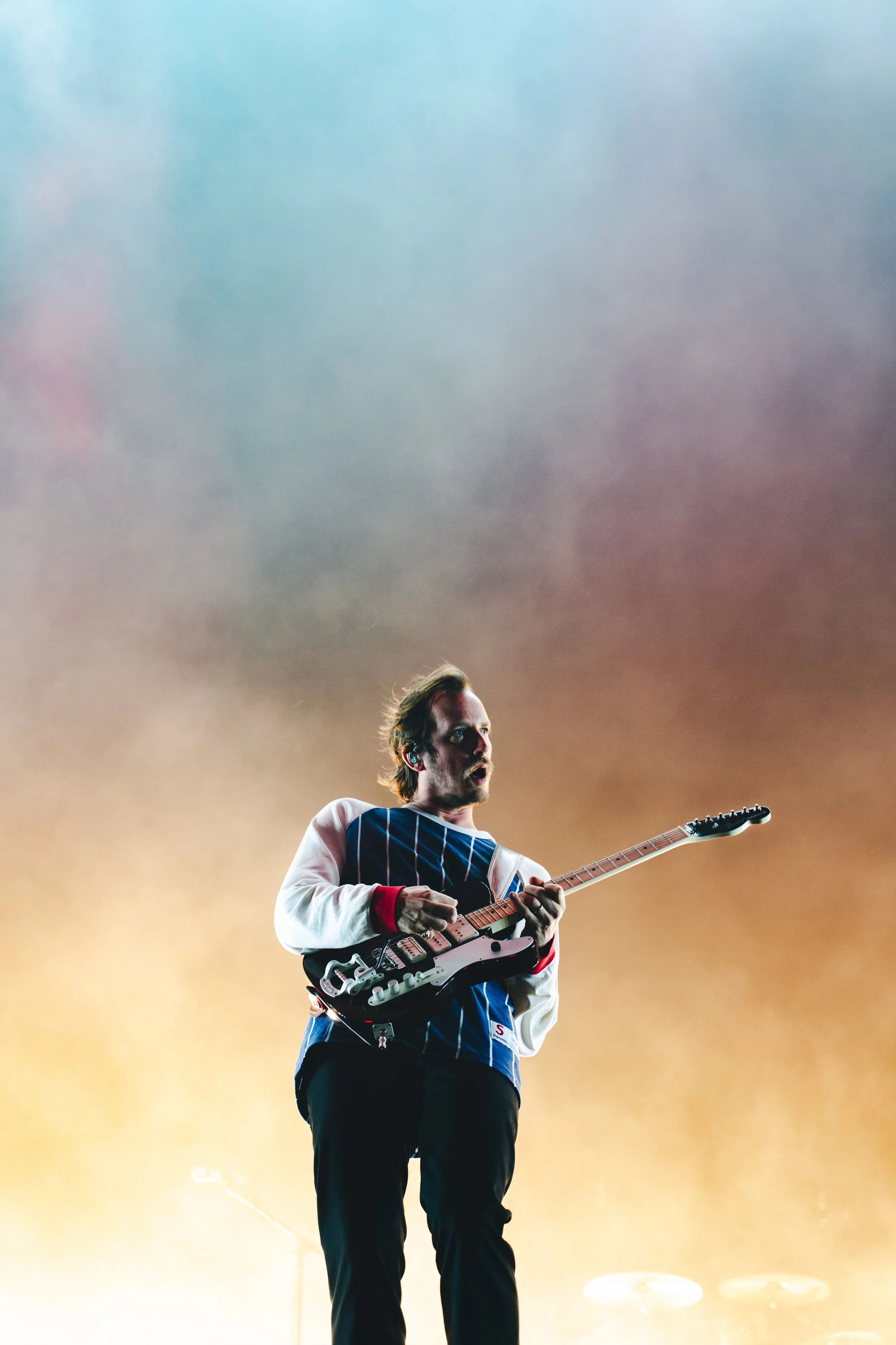 A musician playing an electric guitar on stage with a colorful, cloudy background.