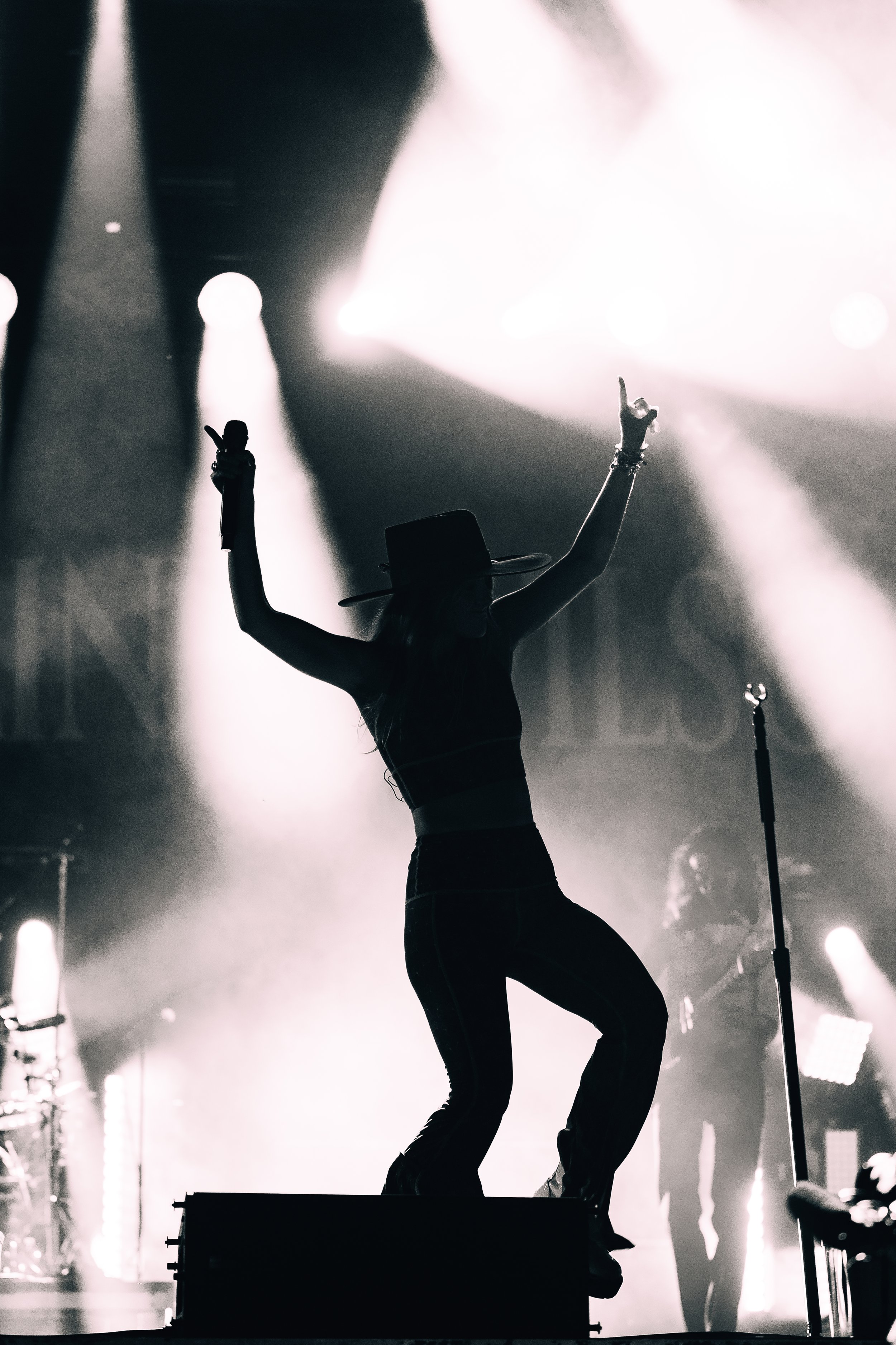Silhouette of a person on stage with arms raised, holding a microphone, illuminated by bright stage lights in black and white.