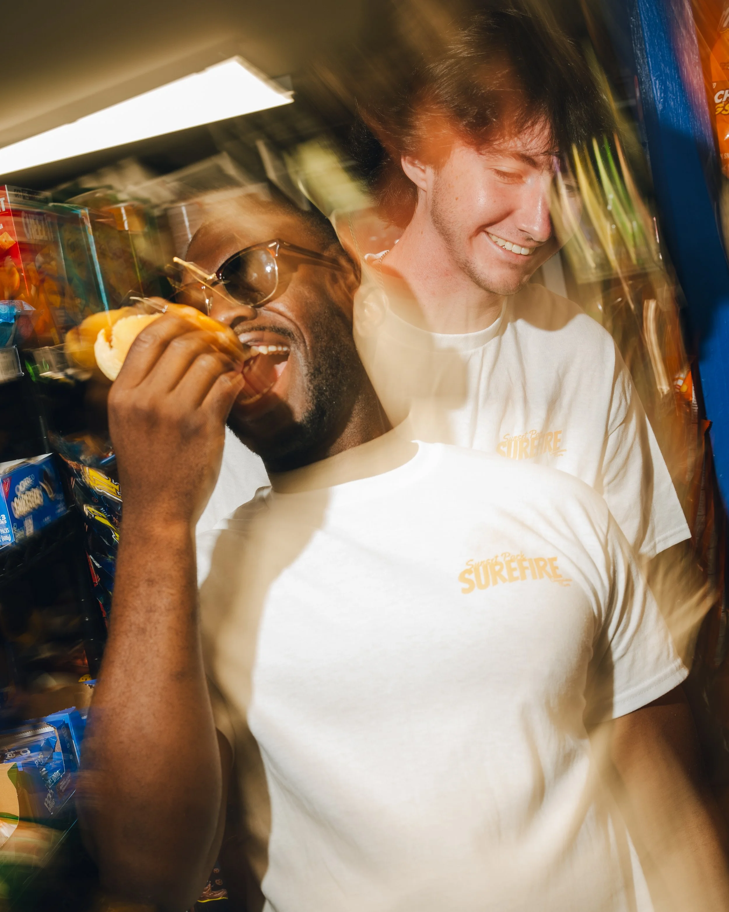 Two young men smiling and enjoying snacks in a grocery store aisle with shelves of various food products.