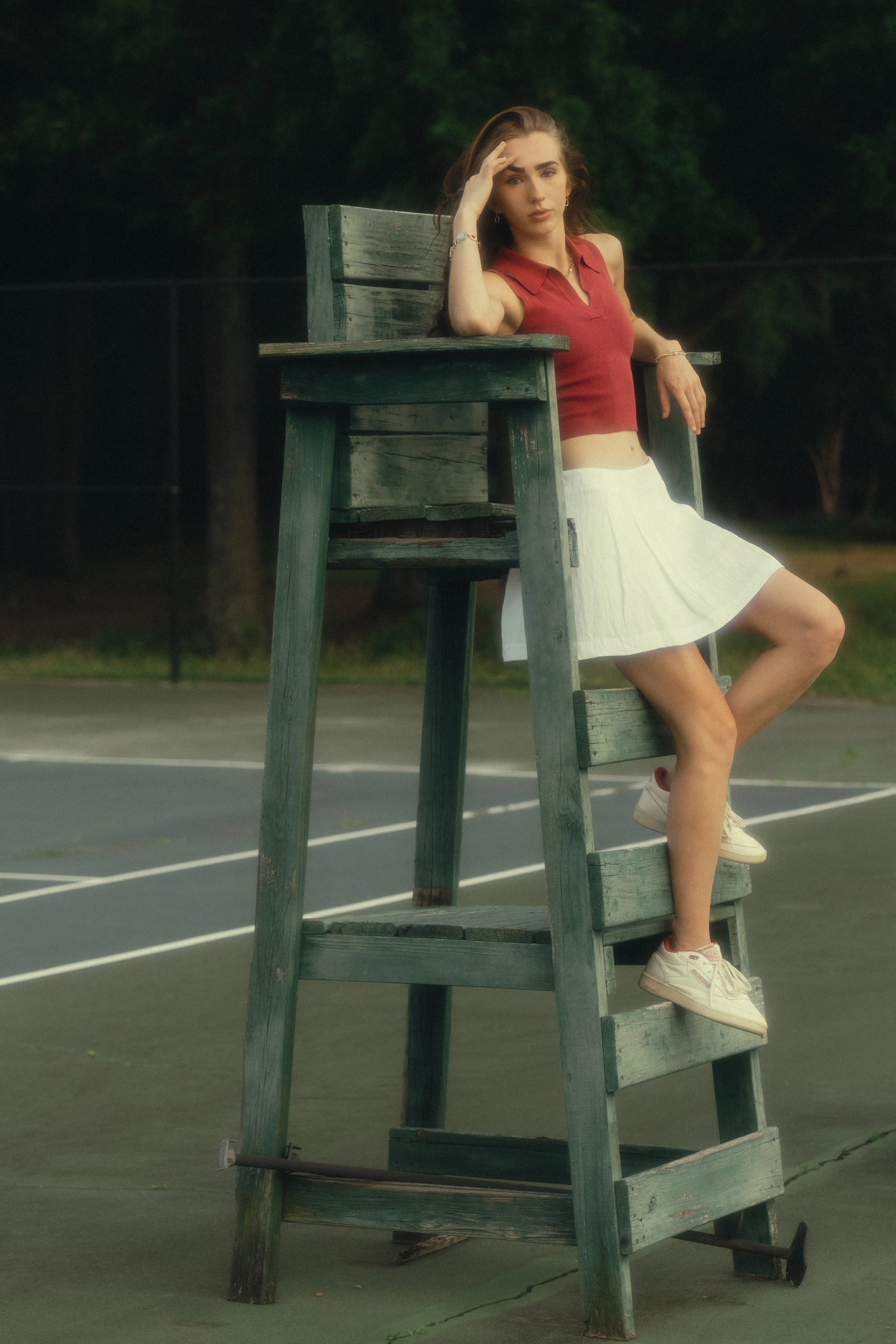 A young woman with long brown hair sitting on an old lifeguard chair on a tennis court, wearing a red sleeveless top, a white skirt, and sneakers, with a contemplative expression, hands on her forehead.