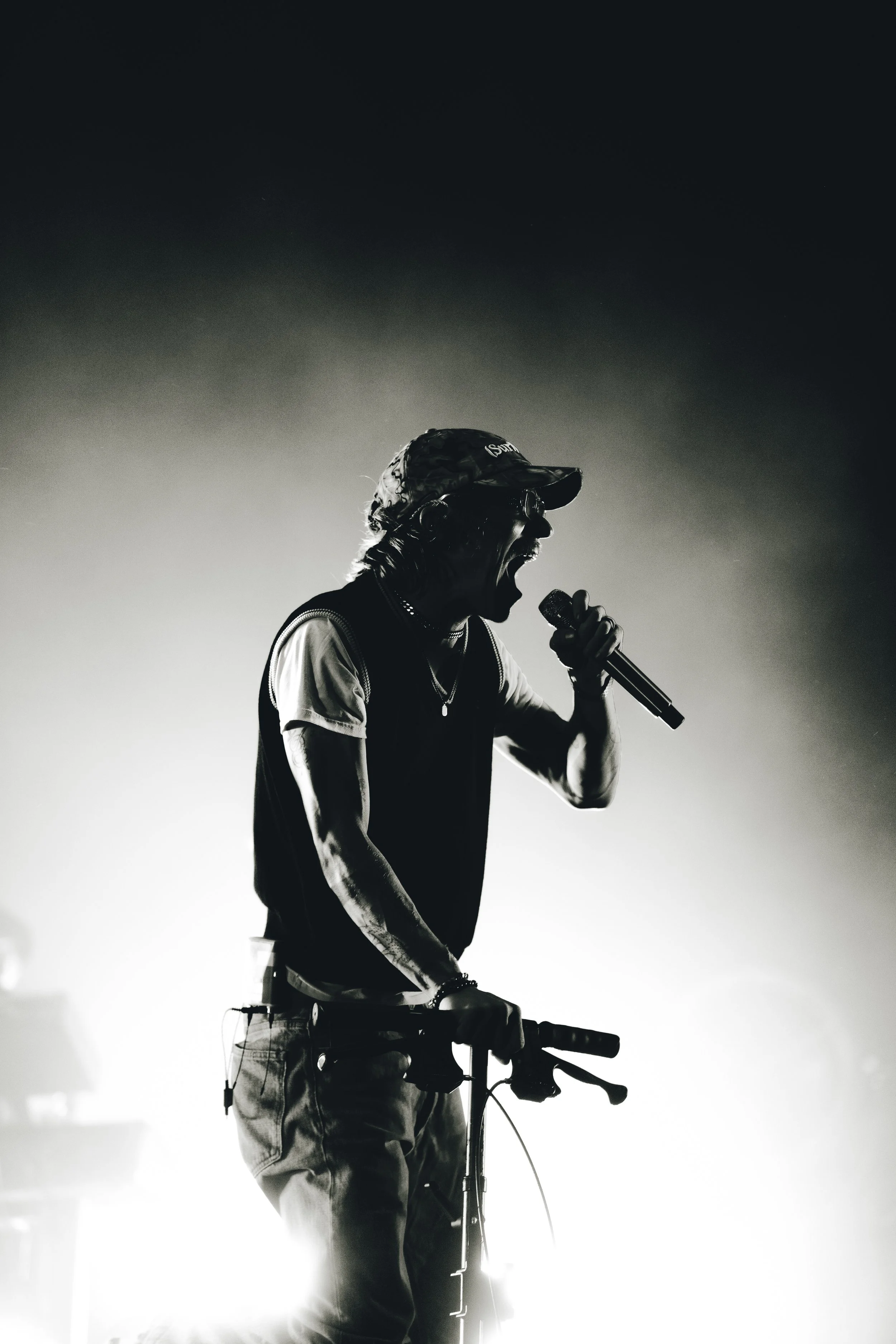 A man wearing a cap and glasses, passionately singing into a microphone while riding a bicycle. The image is in black and white with a dark background.