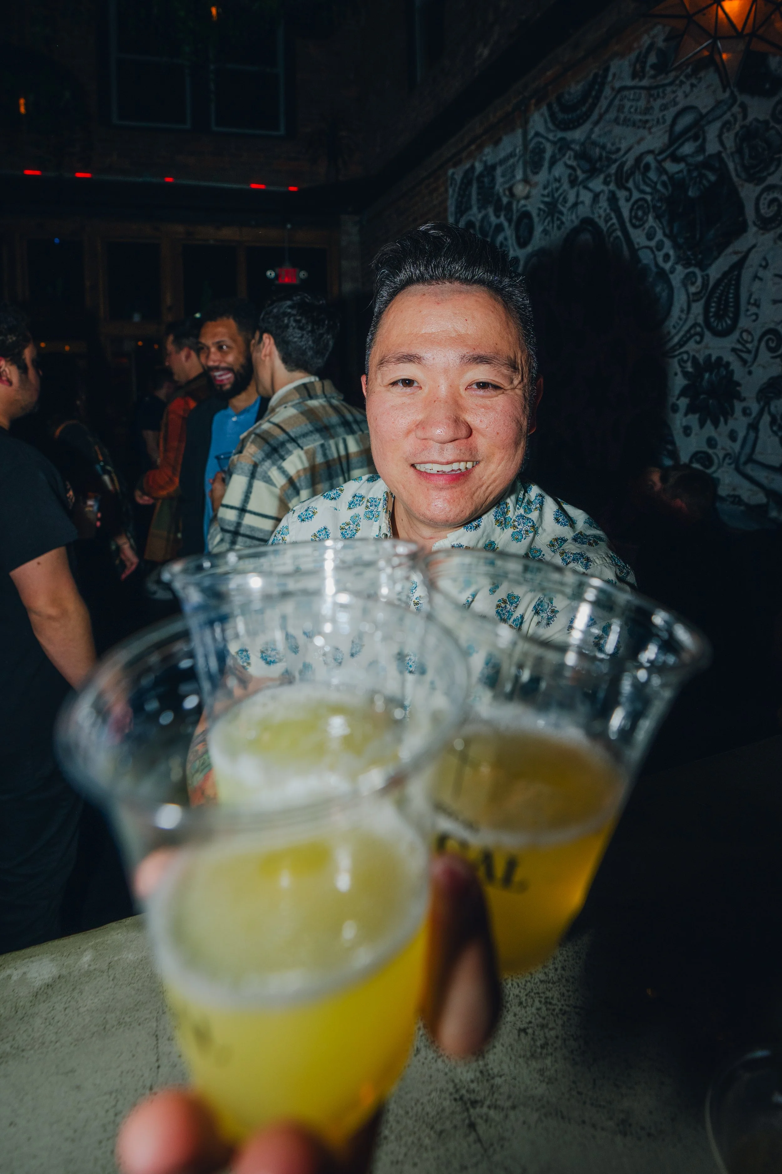 People at a social gathering in a dimly lit venue, holding glasses of yellow-colored drinks with lime slices, with a man smiling at the camera in the foreground, and several other people mingling in the background.