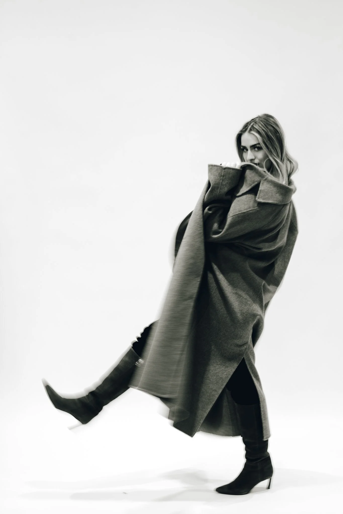 A woman with long, wavy hair wearing a large coat and heeled boots, posing in front of a plain white background.