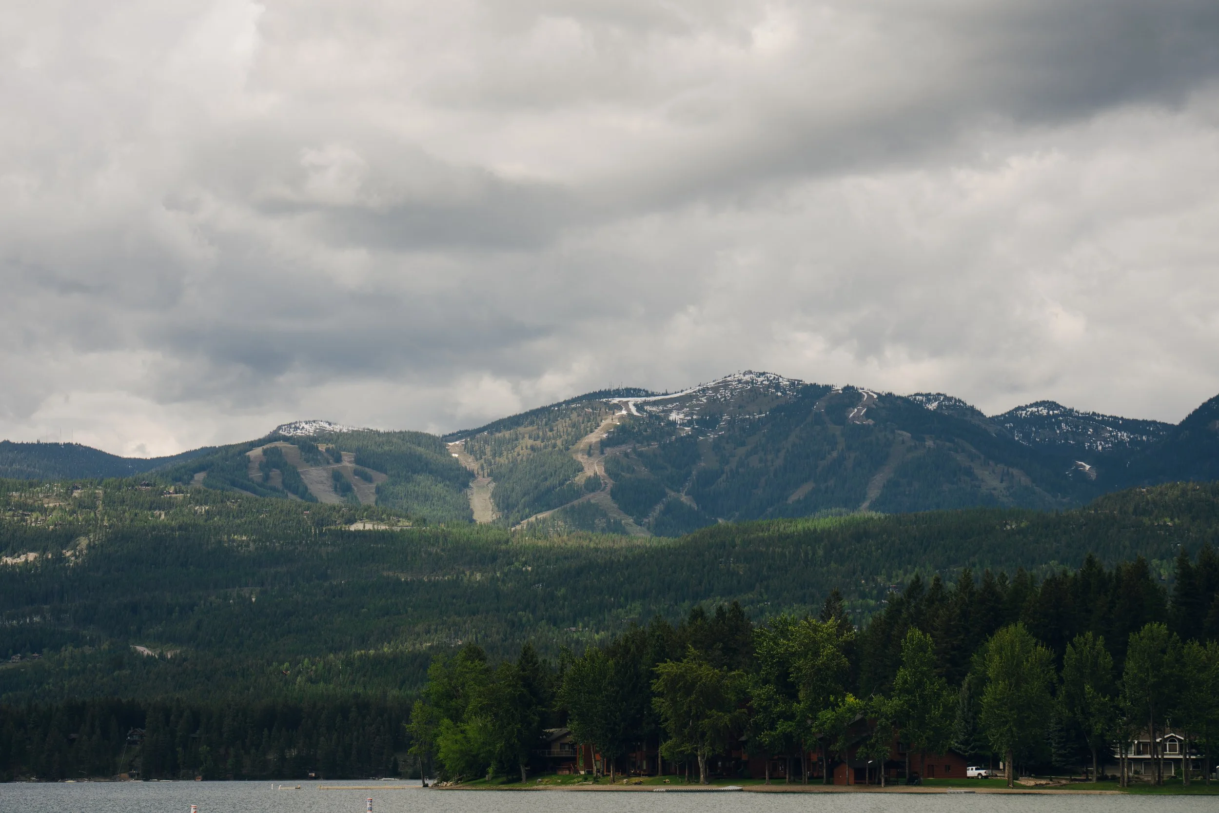 A mountain landscape with forested slopes, snow patches on the peaks, and a cloudy sky overhead. There are buildings near the water in the foreground.