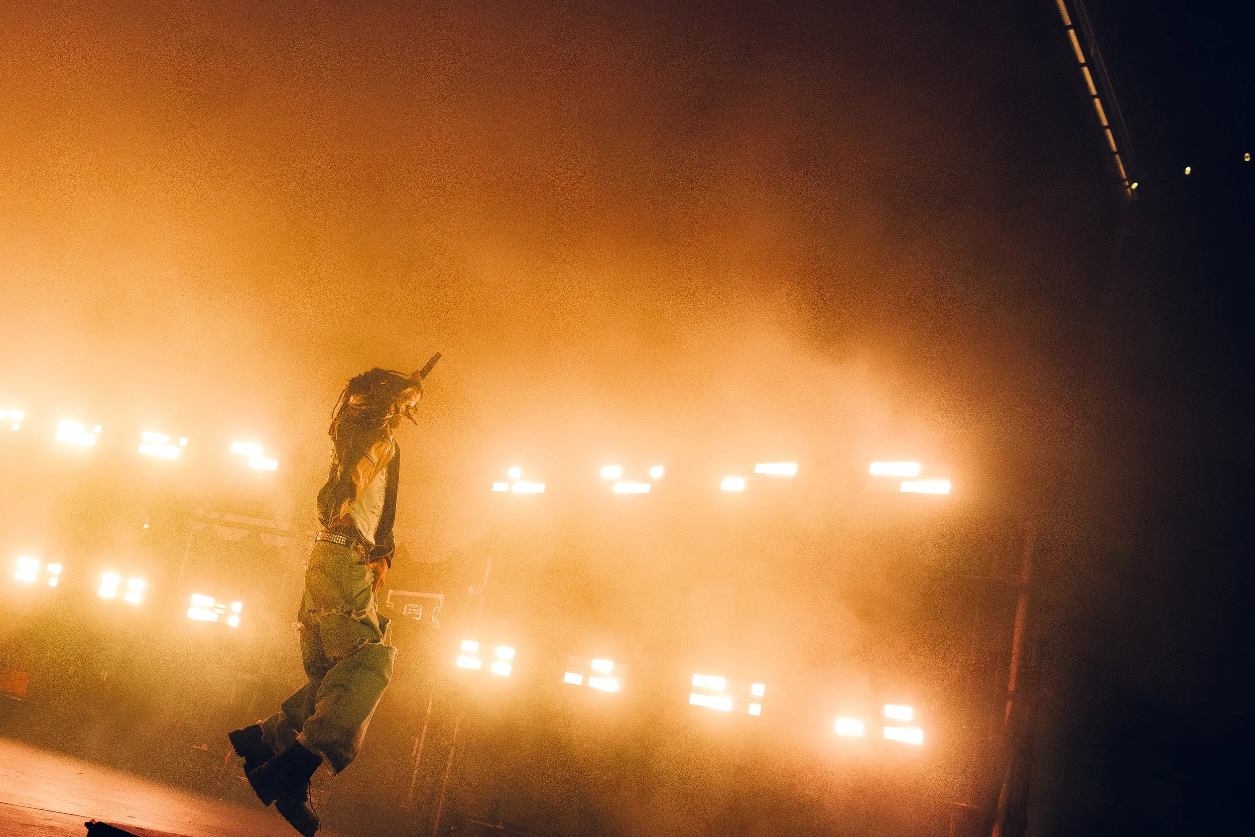 A person with dreadlocks and casual clothing standing on stage with bright lights in the background.