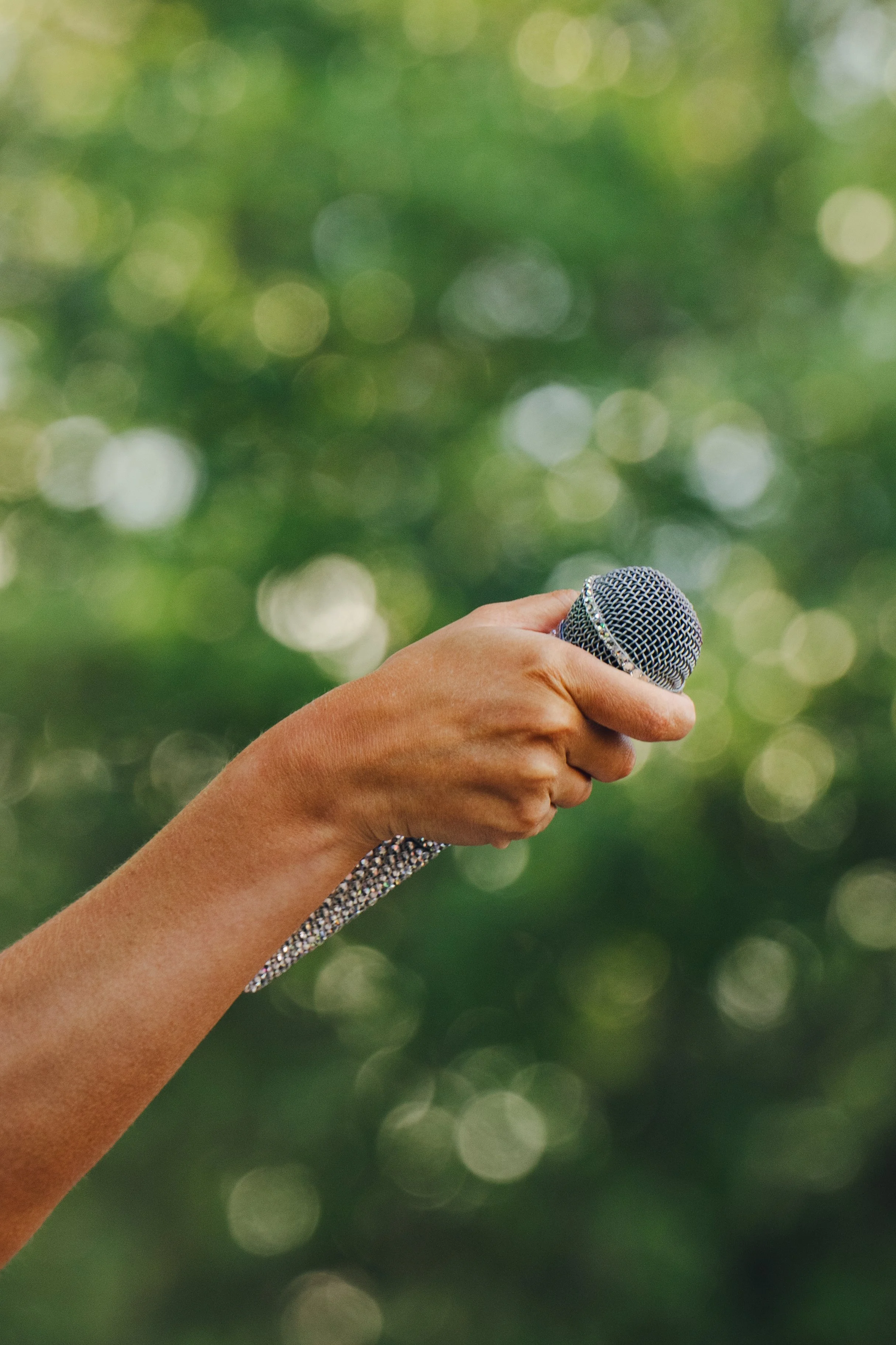A hand holding a microphone with a sparkly strap against a blurred green background.