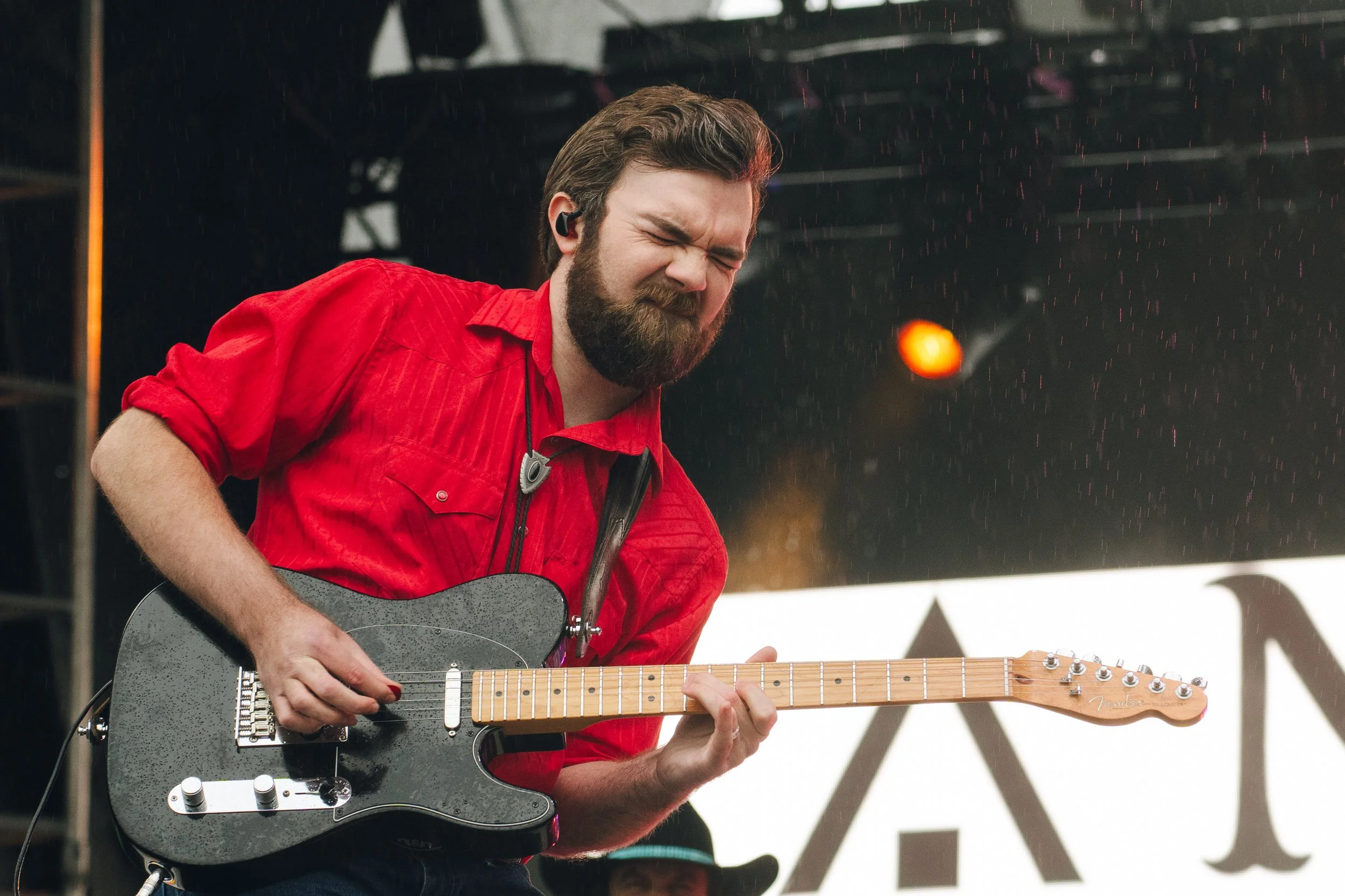 A man with a beard and dark hair, wearing a red shirt, is playing an electric guitar on stage. He appears to be passionate and focused, with rain falling around him.
