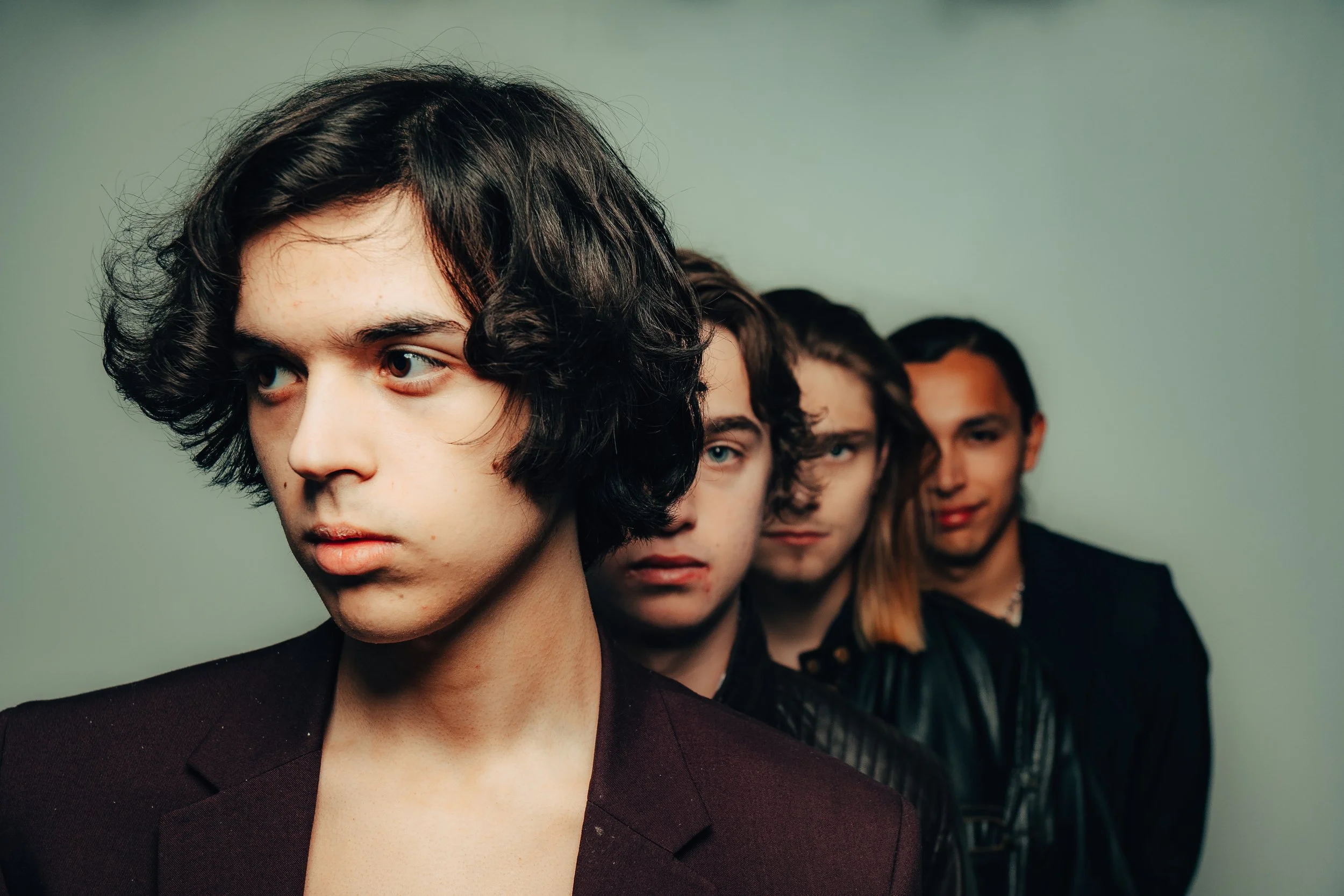Four young adults with dark hair, standing in a line, with serious expressions, against a plain green background.