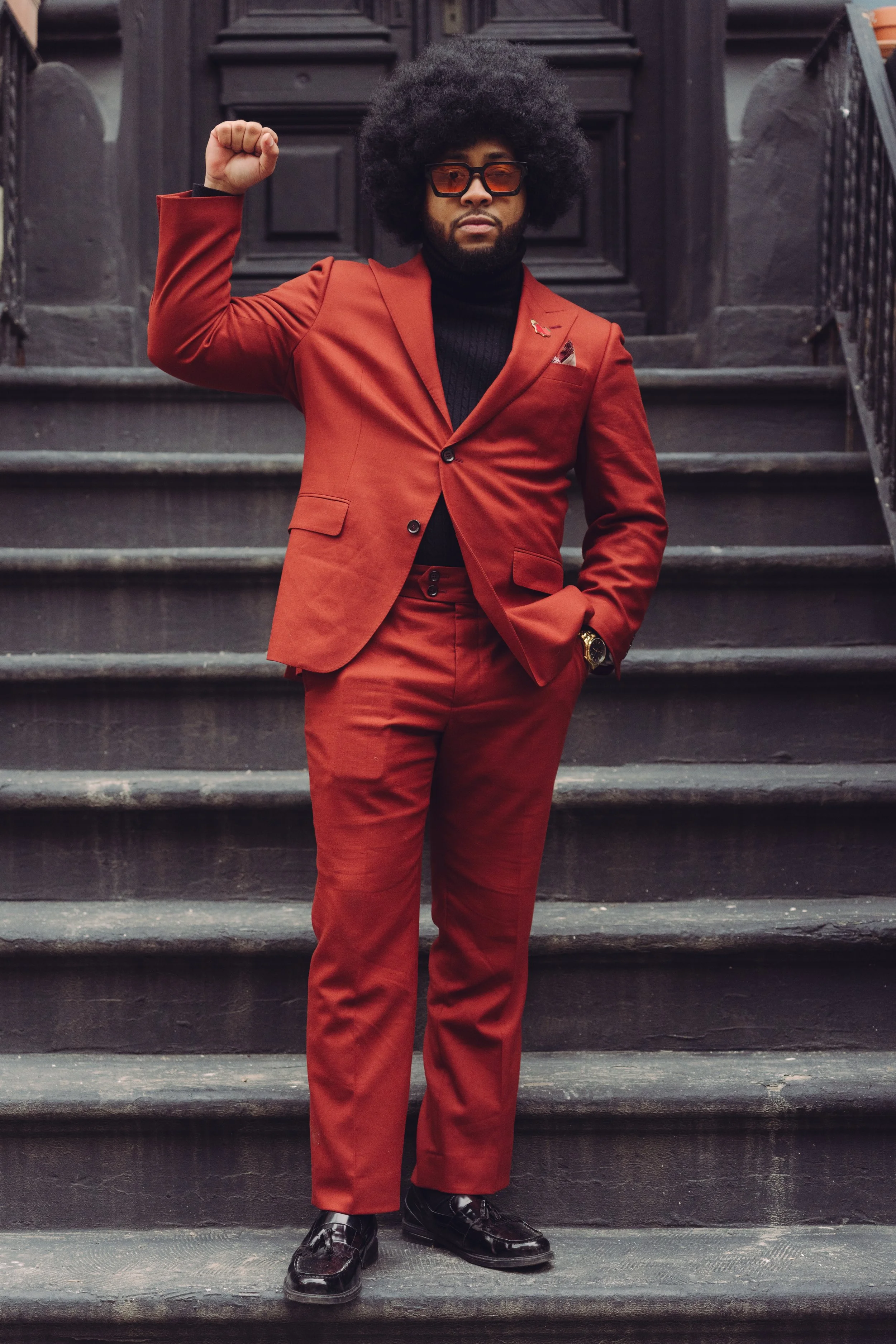A man standing on stairs wearing a red suit, black turtleneck, sunglasses, and shiny black shoes, raising his right fist.