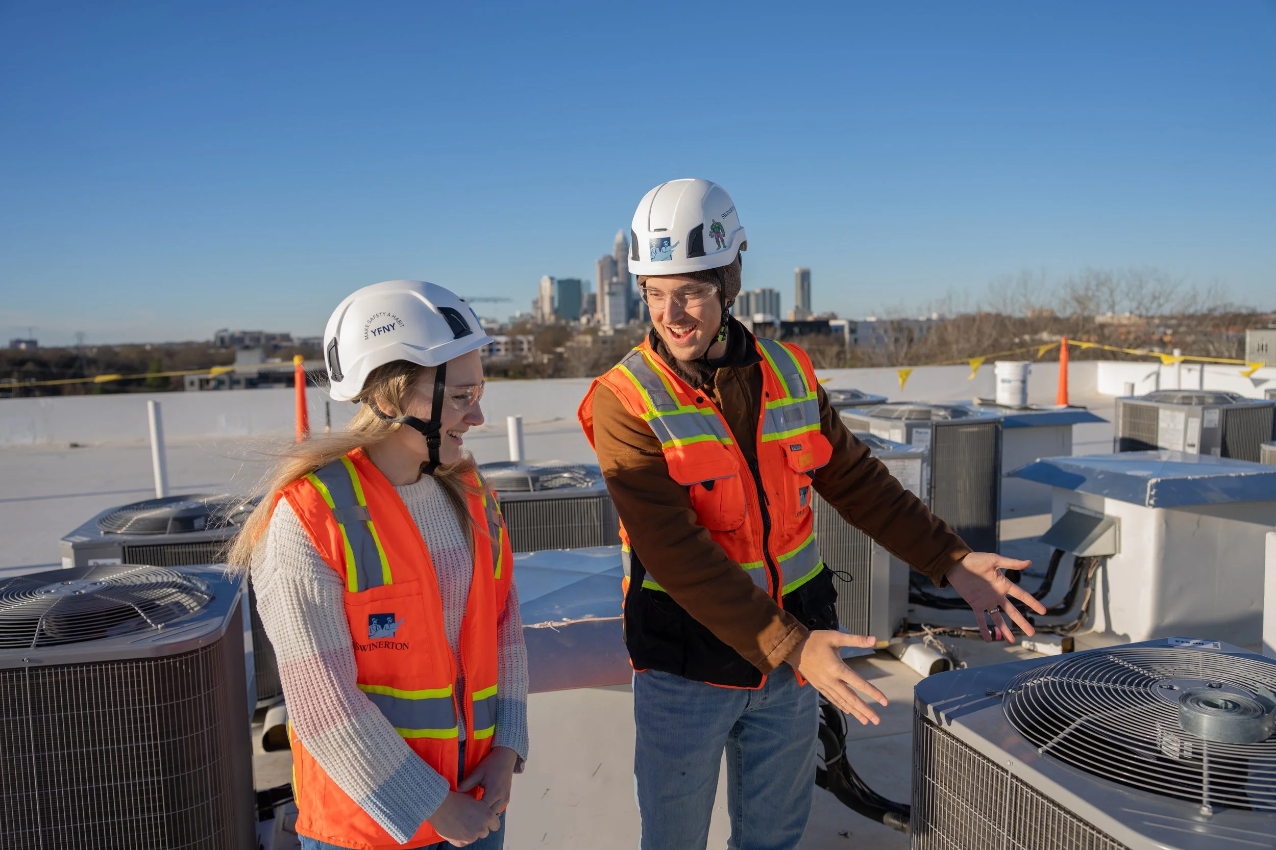 Two people, a man and a woman, wearing orange safety vests and white helmets standing on a rooftop with HVAC air conditioning units, city skyline in the background, clear blue sky.