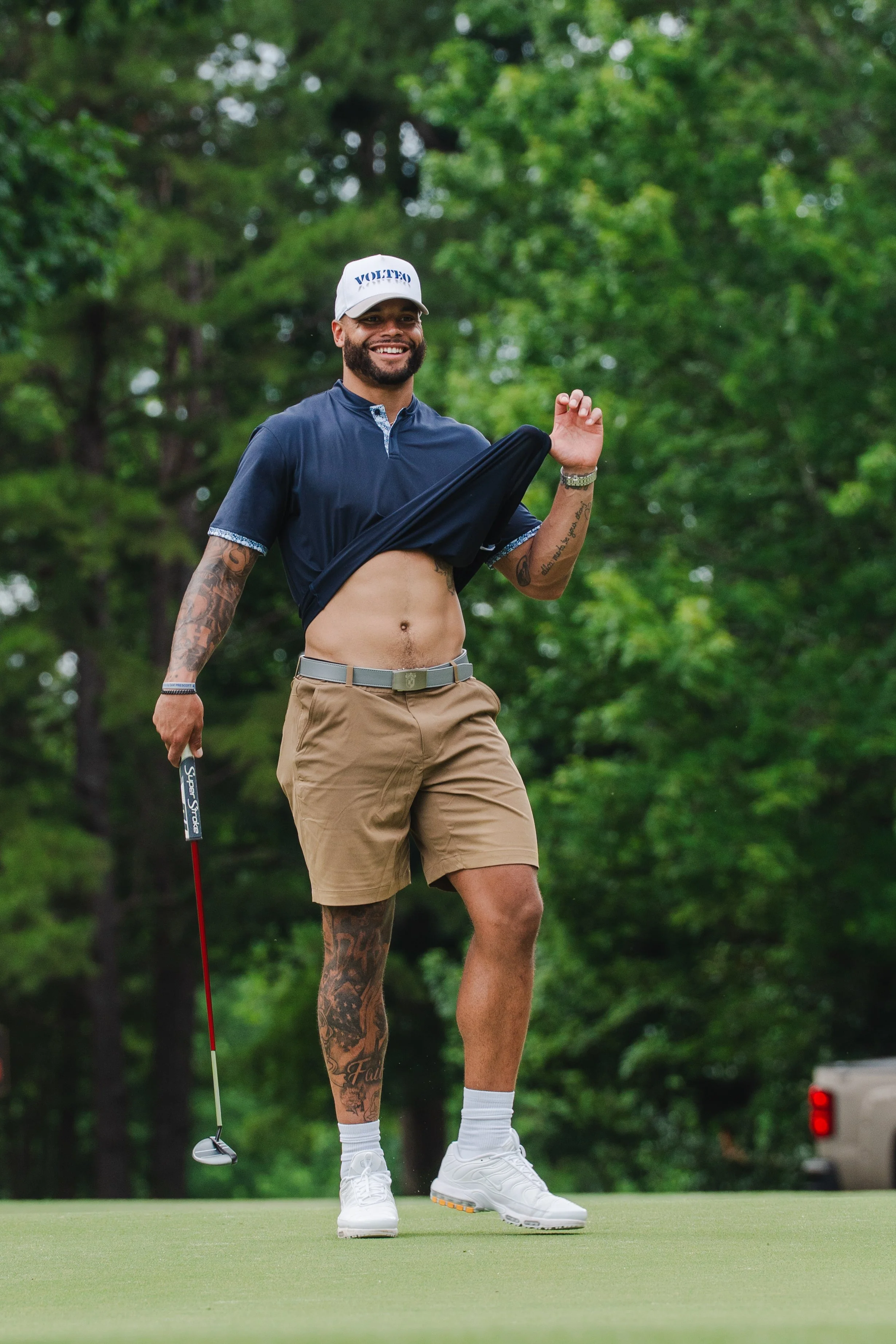 A man on a golf course wearing tan shorts, a navy blue shirt raised to reveal his abdomen, white sneakers, and a white cap that says 'VOTED.' He is smiling and holding a golf club, with a vibrant green background of trees.
