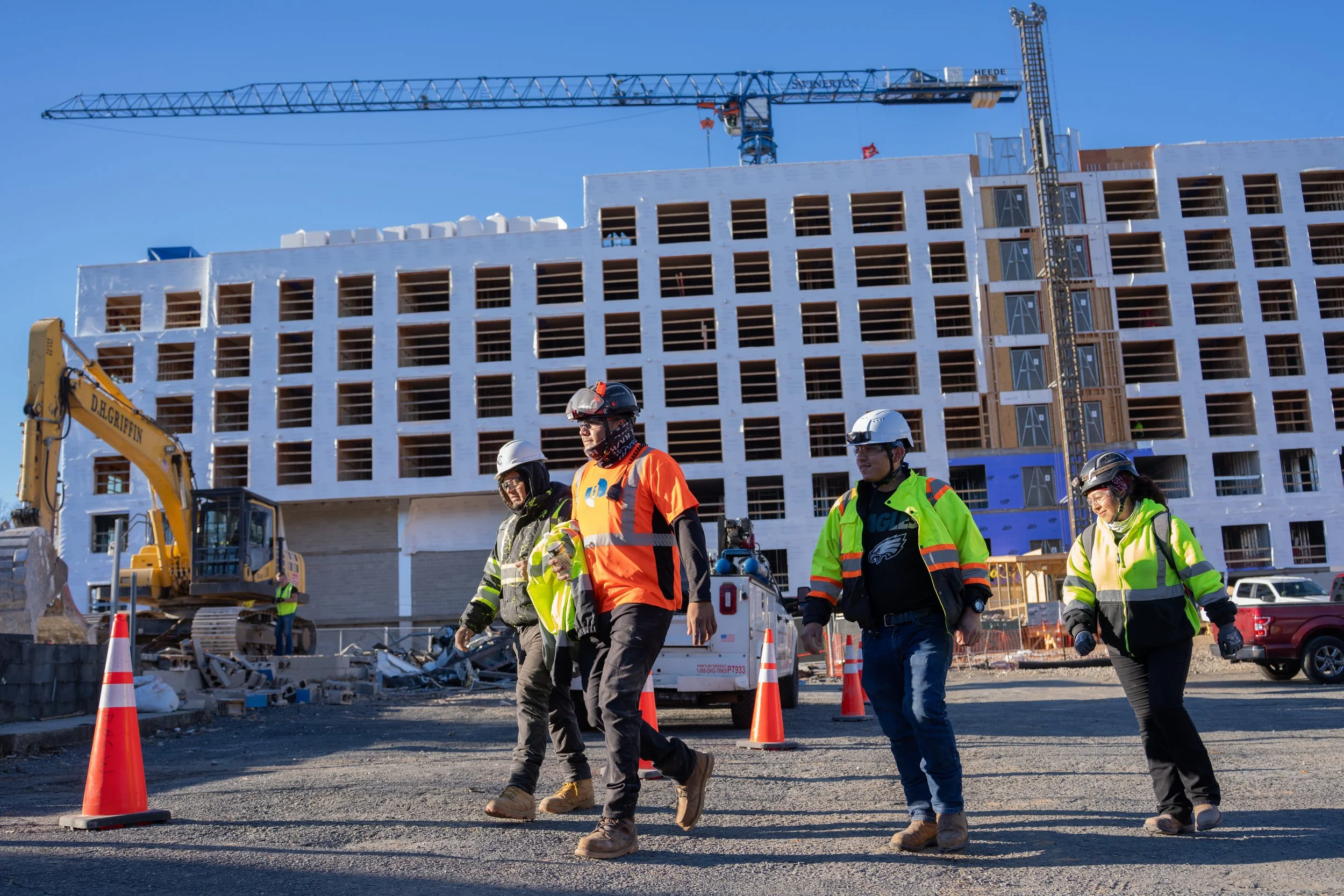Construction workers wearing safety gear walk on a construction site in front of a building under construction and a crane.
