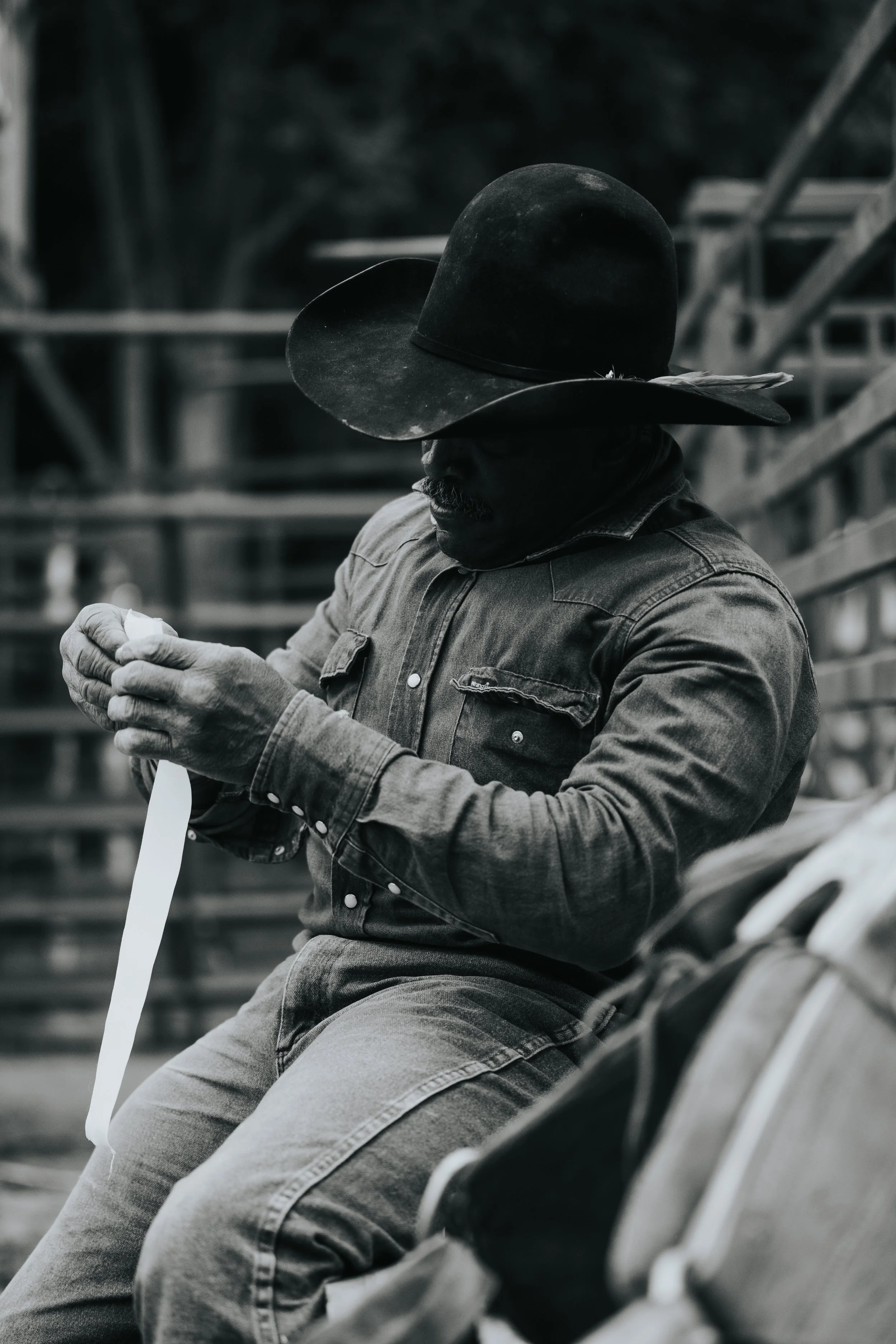 A man sitting on a bench, wearing a large brimmed hat, using his phone, in black and white.