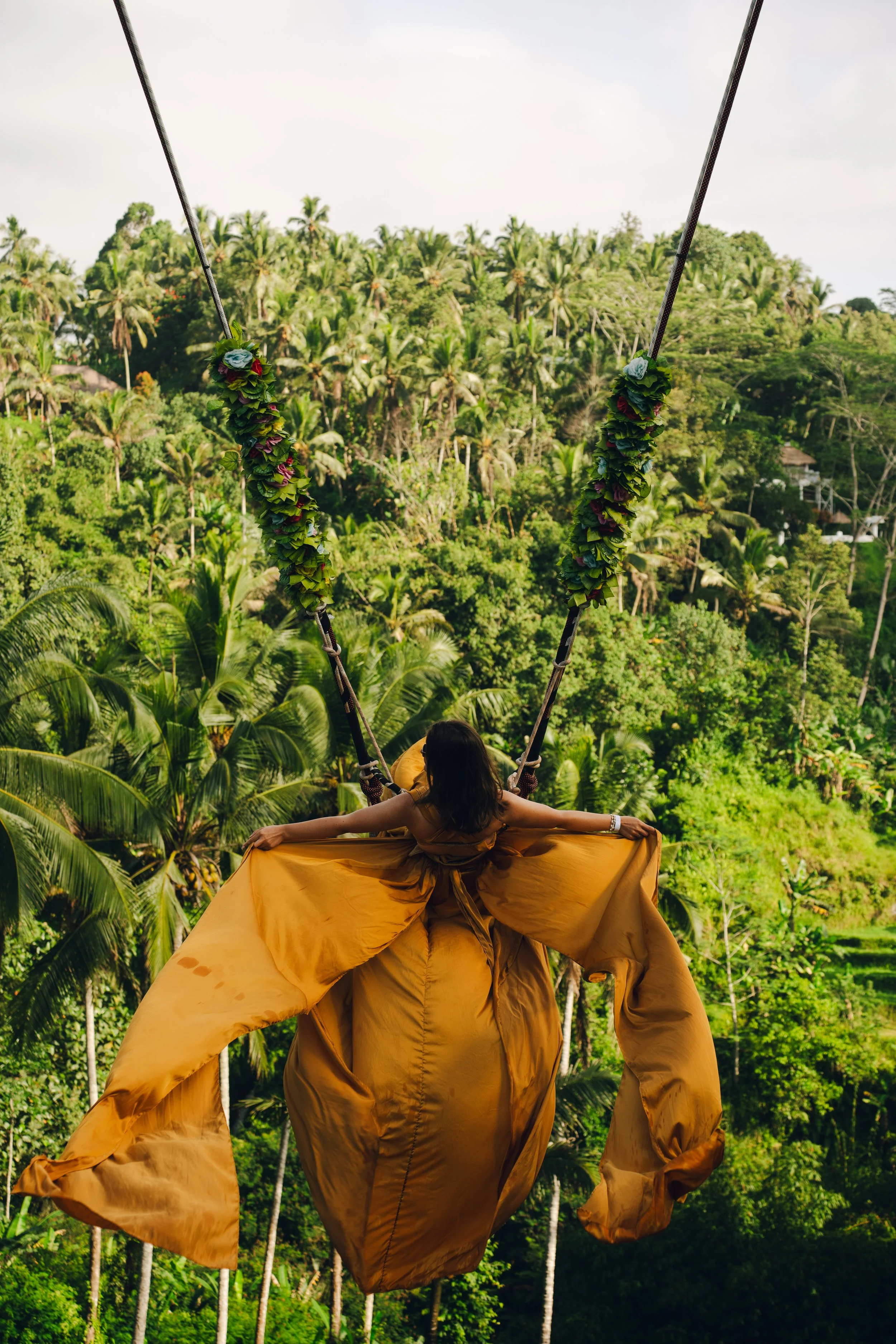 A woman in a flowing golden dress with a full skirt is sitting on a swing decorated with flower garlands over a lush green tropical forest.