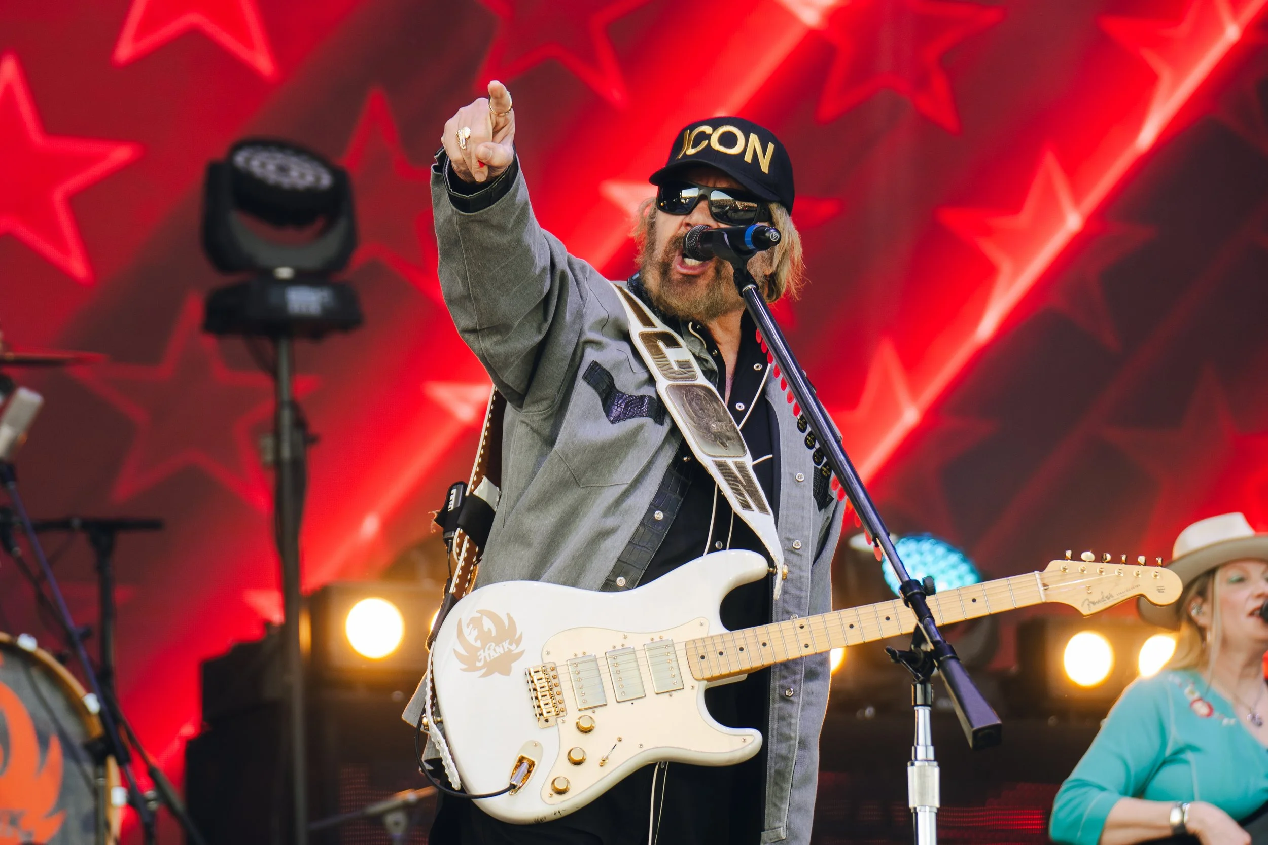 A man with long blond hair wearing sunglasses, a black cap with gold lettering, and a gray jacket is singing into a microphone on a stage. He is playing a white electric guitar with golden hardware. The stage background has large red star decorations