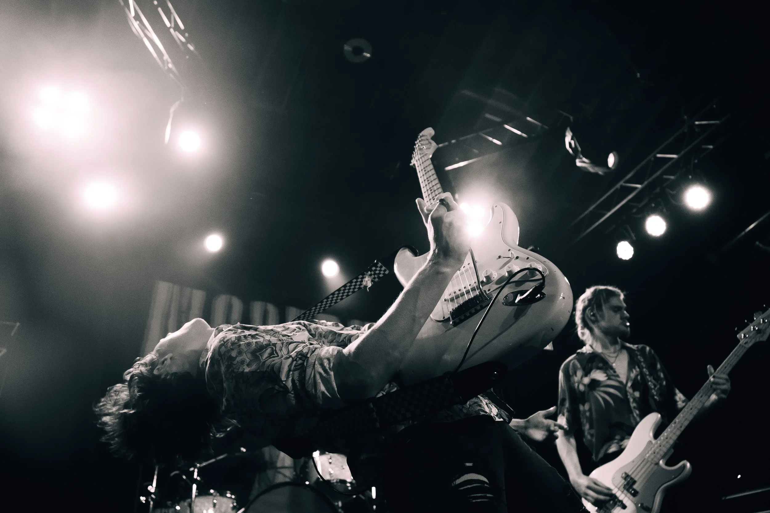 Musicians performing on stage, with one person lying on the ground playing an electric guitar and another standing and playing a bass guitar, under bright stage lights in black and white.