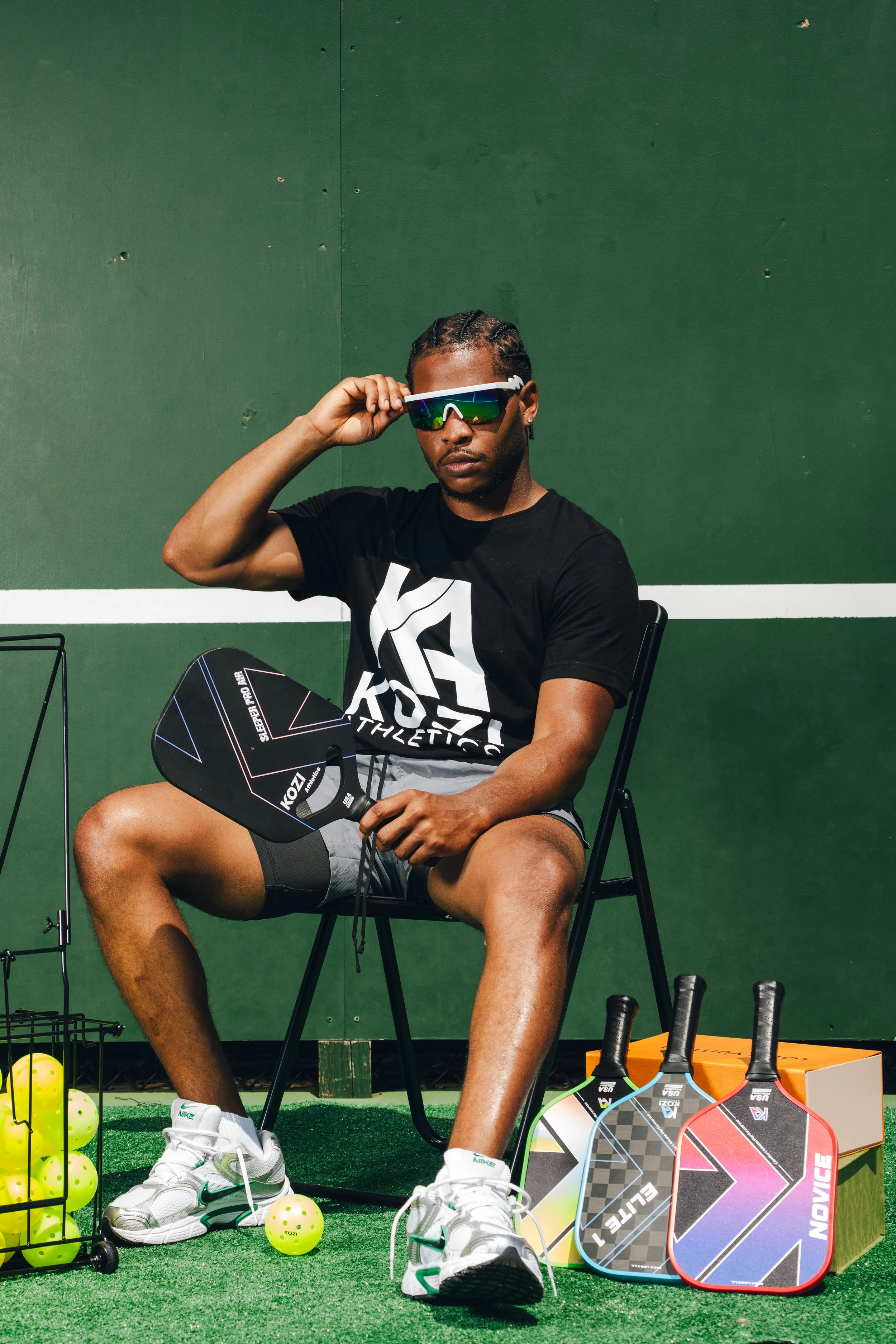 A man sitting on a black chair outdoors, holding a pickleball paddle, wearing sunglasses, a black T-shirt, gray shorts, and white sneakers. There is a stack of multiple pickleball paddles, a box, tennis balls, and tennis rackets nearby on artificial 