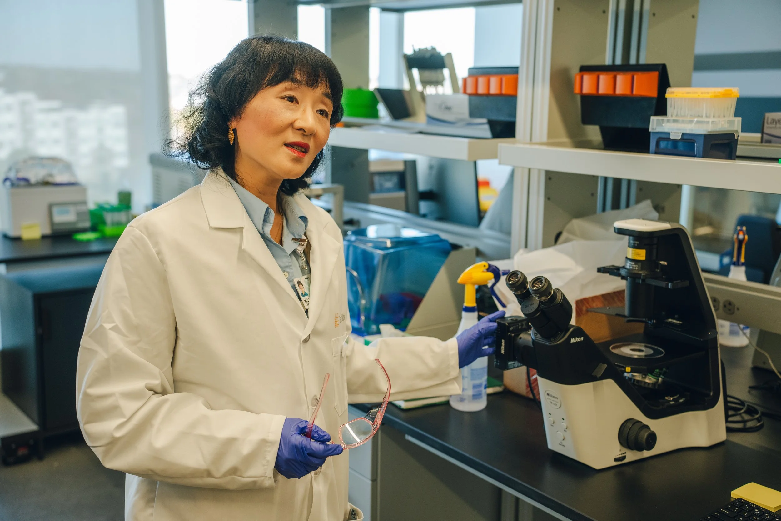 A scientist in a laboratory wearing a white coat and purple gloves, holding safety glasses, standing next to a stereo microscope. The lab has shelves with lab supplies and equipment.