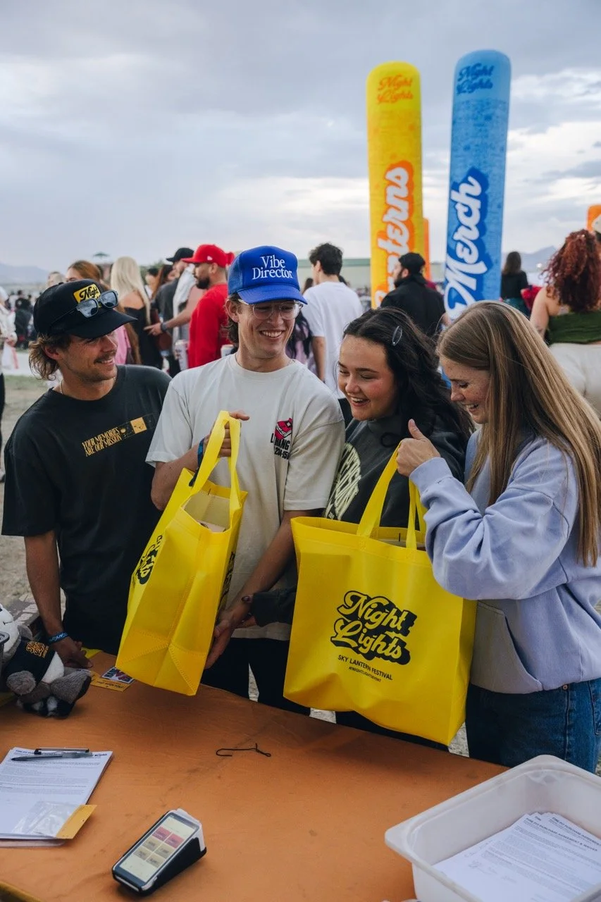 Four people smiling and holding yellow tote bags at an outdoor festival, with large colorful banners and other attendees in the background.