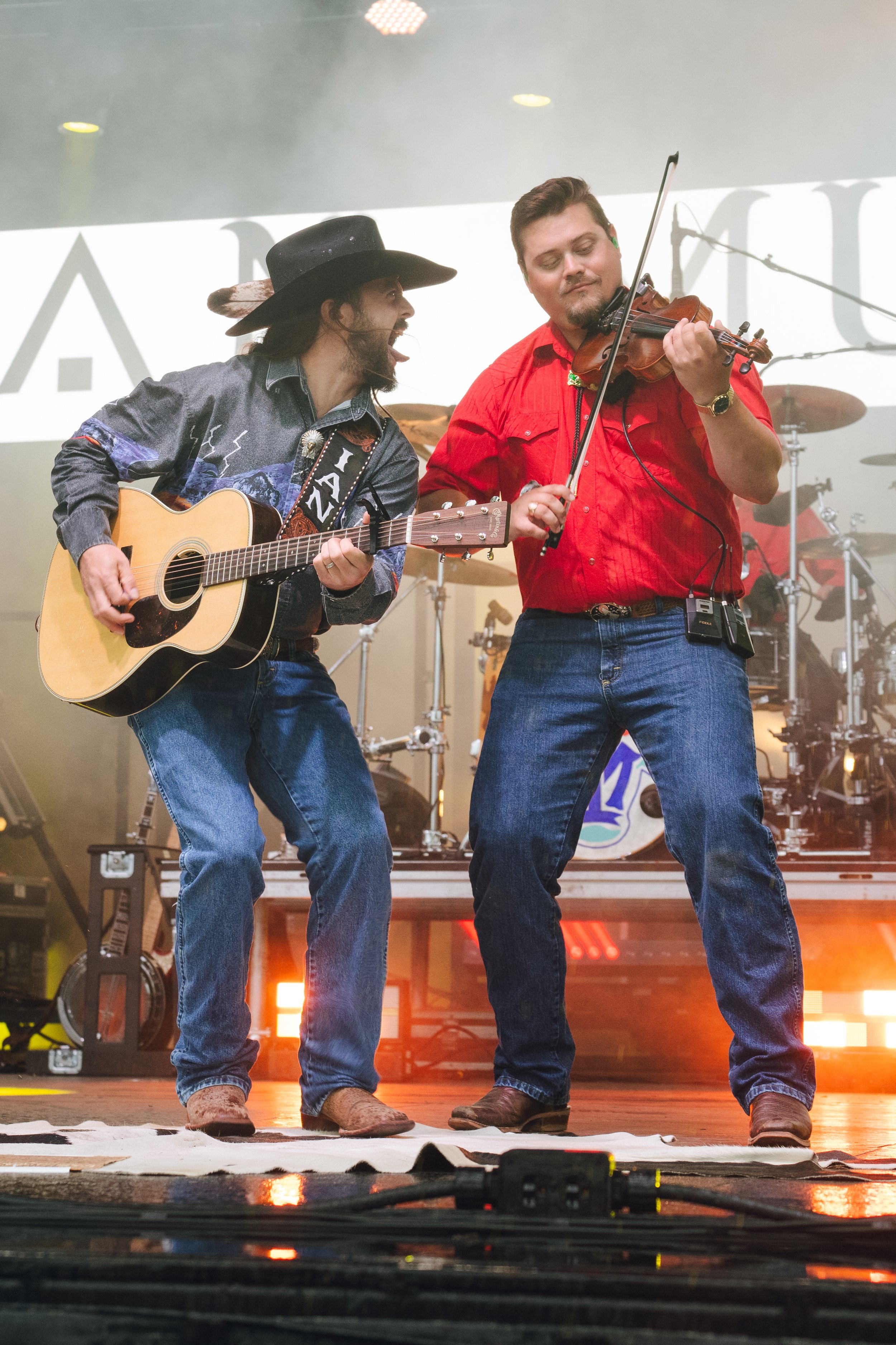 Two musicians performing on stage; one playing the acoustic guitar and singing, the other playing the violin, both dressed casually, with band equipment and drums visible in the background.