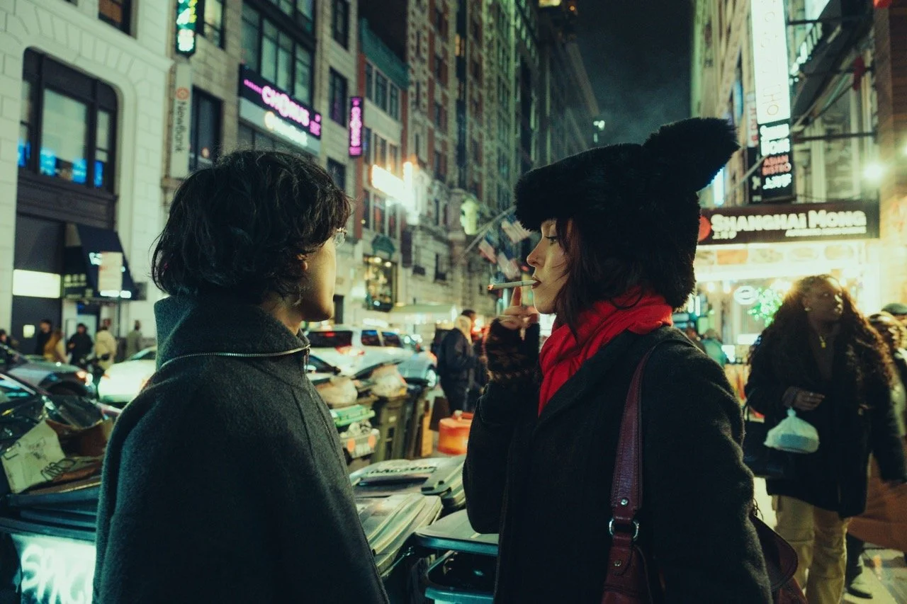 Two women stand on a busy city street at night, facing each other, one smoking a cigarette. The woman on the left has short curly hair and wears a dark coat. The woman on the right has medium-length dark hair, wears a large black fuzzy hat, a red sca