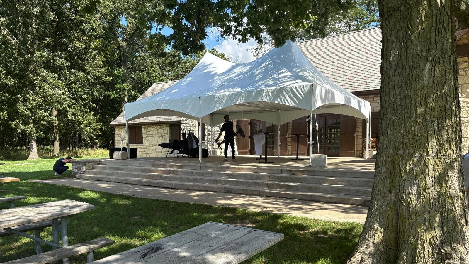 Stage set up outdoors under a white canopy with a person standing near musical instruments, surrounded by trees and a building in the background.
