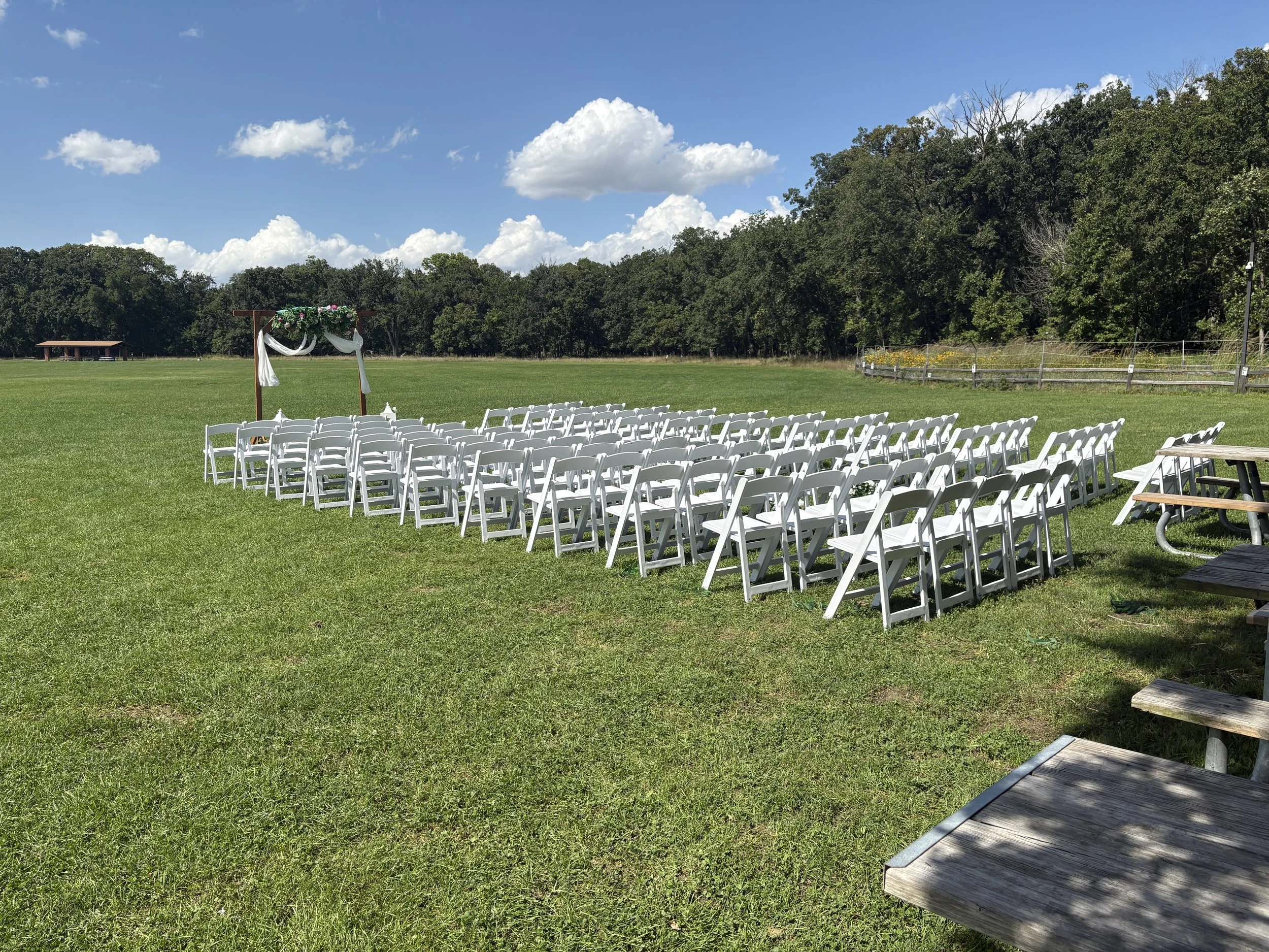 Outdoor wedding setup with white chairs arranged in rows facing a decorated arch on a grassy field under a blue sky with clouds, surrounded by trees and nature.