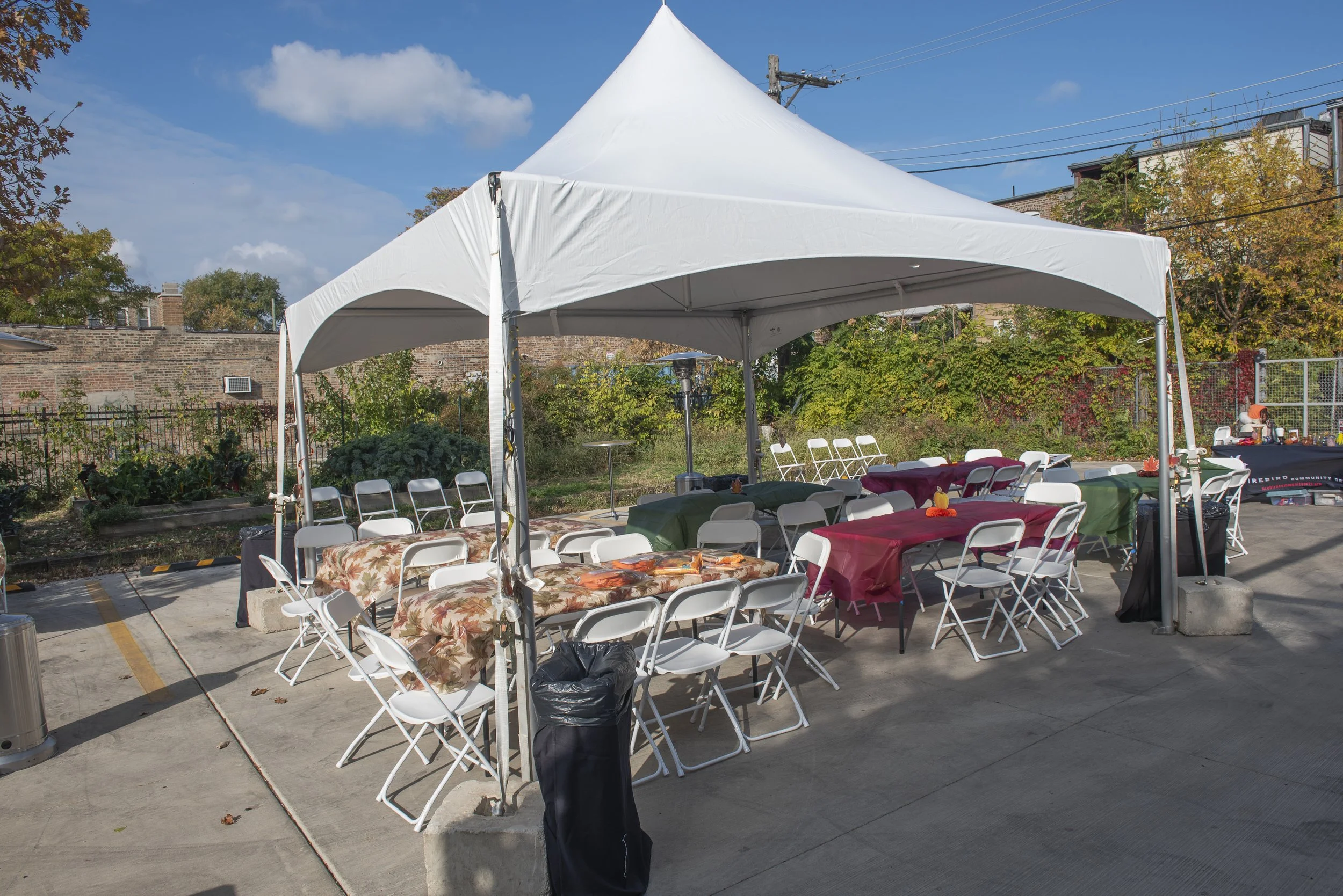 An outdoor event setup with a large white canopy tent, decorated tables covered with red and green tablecloths, and white folding chairs. There are some orange fall-themed decorations on the tables, and a background of trees, a brick wall, and a clear blue sky.