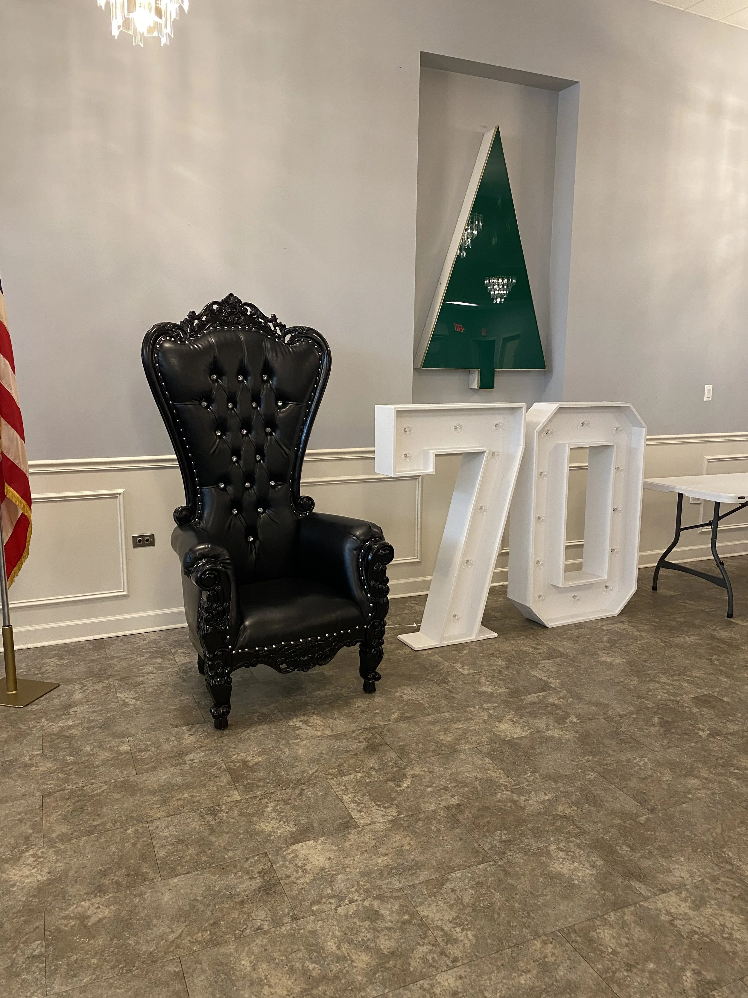 A black, high-backed, ornate leather chair with Victorian-style carving and button-tufted upholstery, is placed next to decorative white marquee letters '7' and '0' in an interior space with gray walls and brown floor tiles, with an American flag partially visible on the left and a modern triangle-shaped mirror on the wall behind.