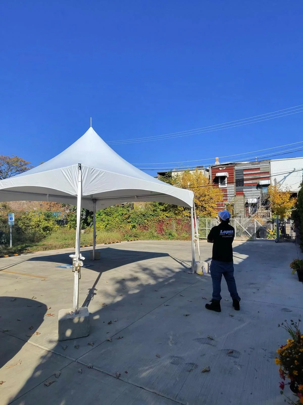 A person standing under a white event tent in a parking lot, with buildings and autumn trees in the background and power lines overhead.