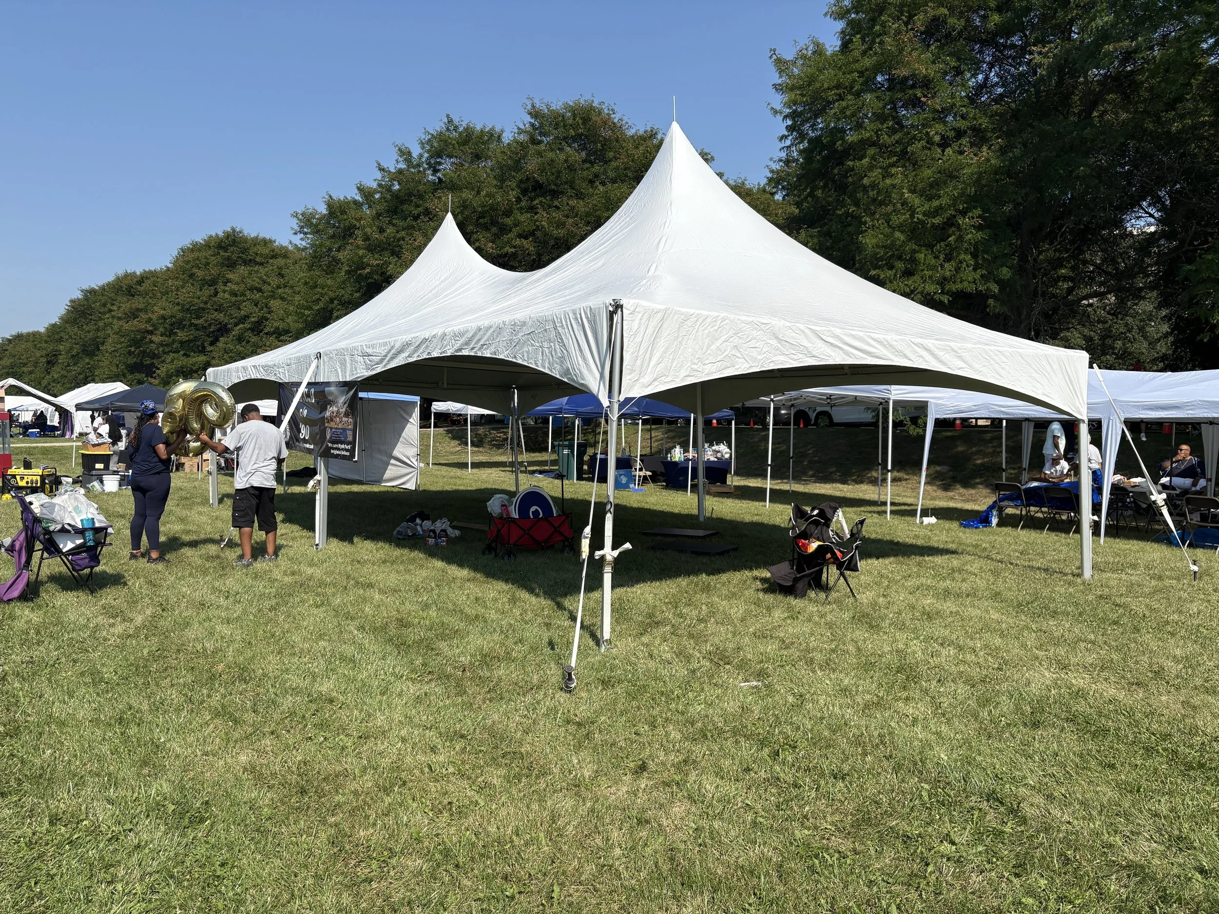 A large white canopy tent set up outdoors with people and equipment underneath, in a grassy area with trees in the background.