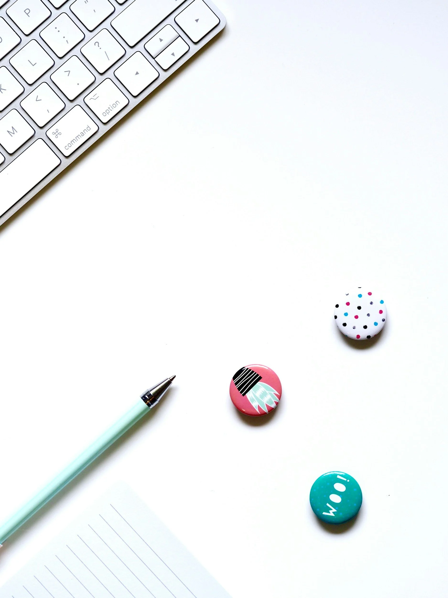 Minimalist workspace with a white keyboard, colorful pins, light blue pen, and a lined notepad on a white desk.