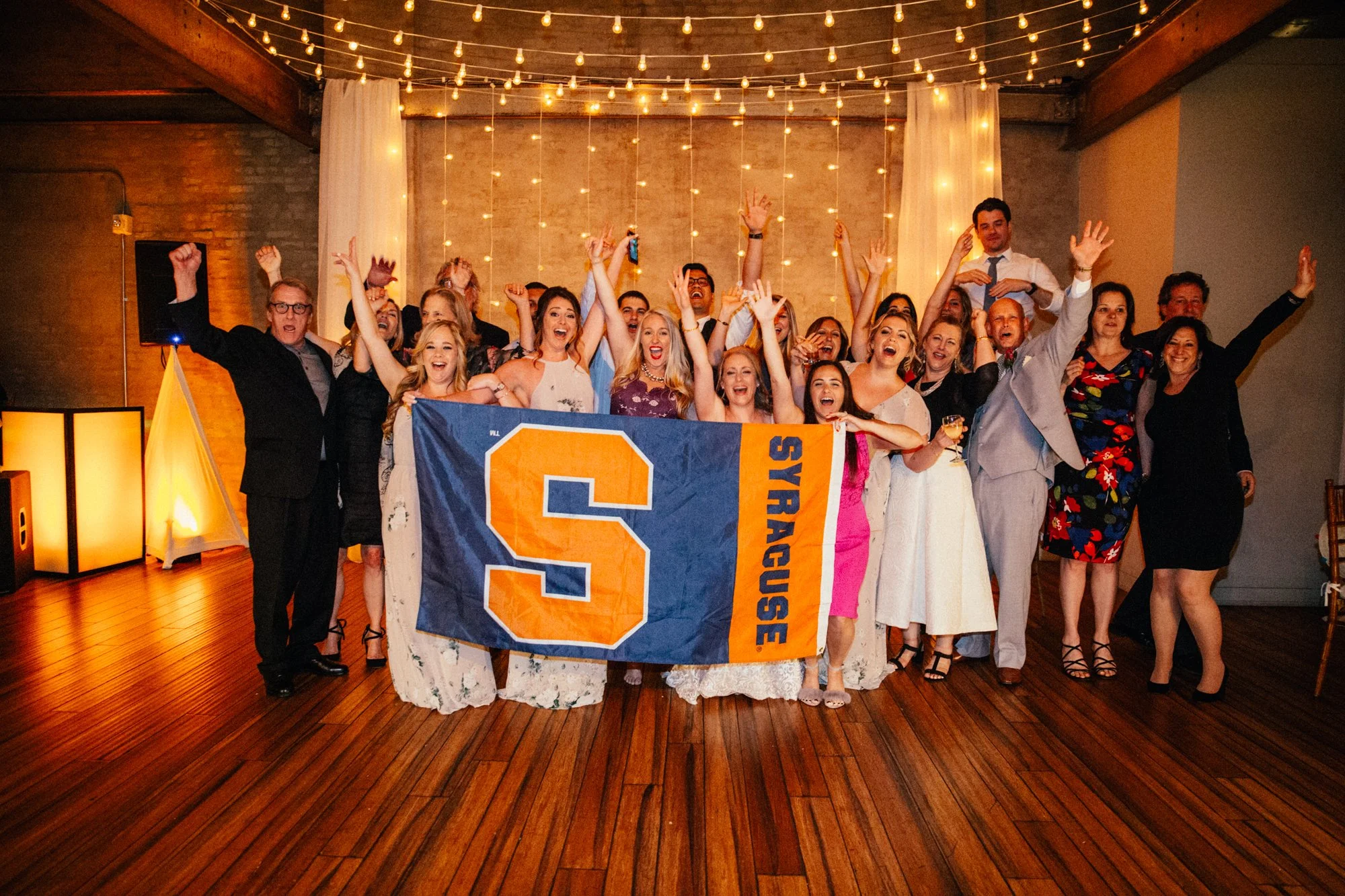 Group of people celebrating during a wedding reception, holding a Syracuse University flag, with festive lights hanging above.