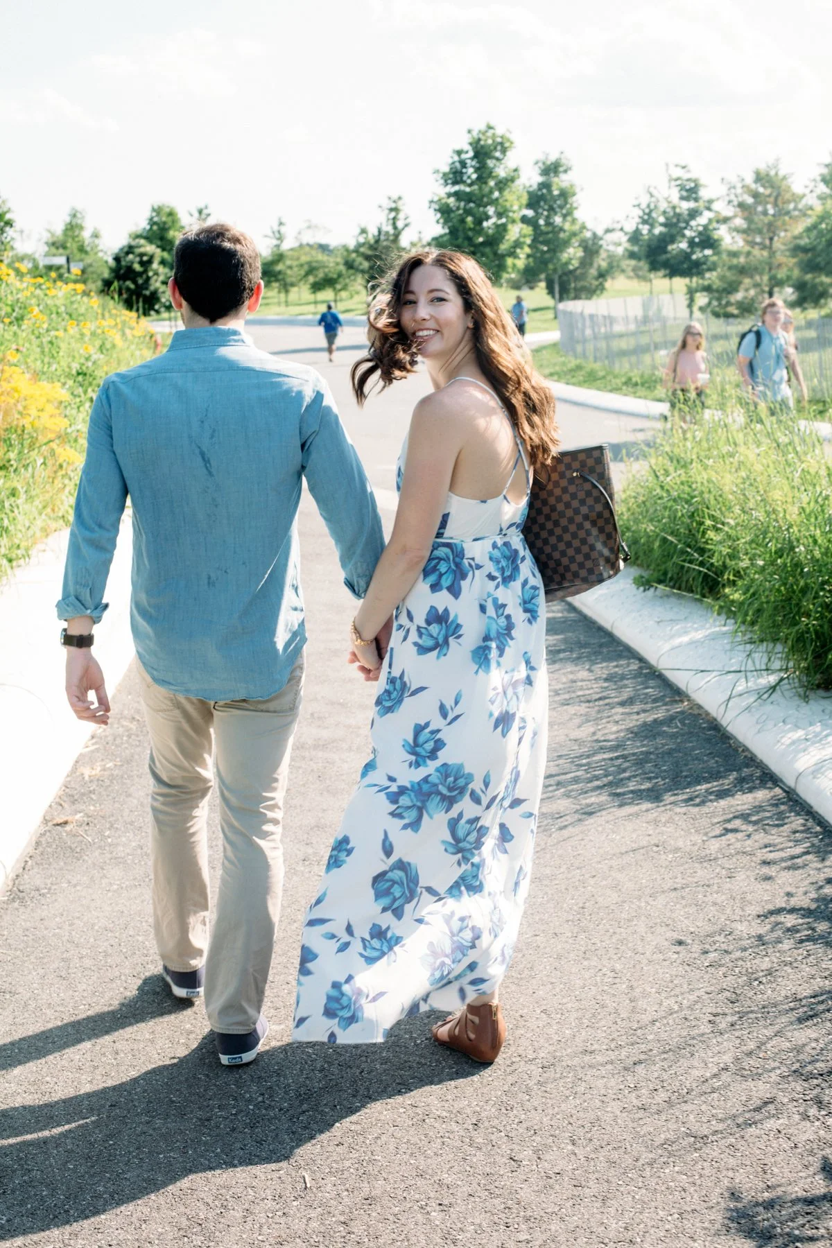 A couple walking hand in hand on a sunny pathway, with the woman wearing a white dress with blue floral patterns and carrying a bag, while the man wears a light blue shirt and beige pants. Trees and grass are visible along the path.