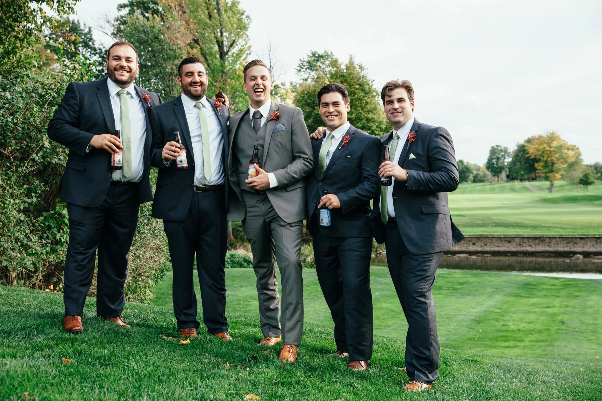 Groom and groomsmen in formal suits smiling, standing outdoors on a grassy area with trees in the background, holding beer bottles.