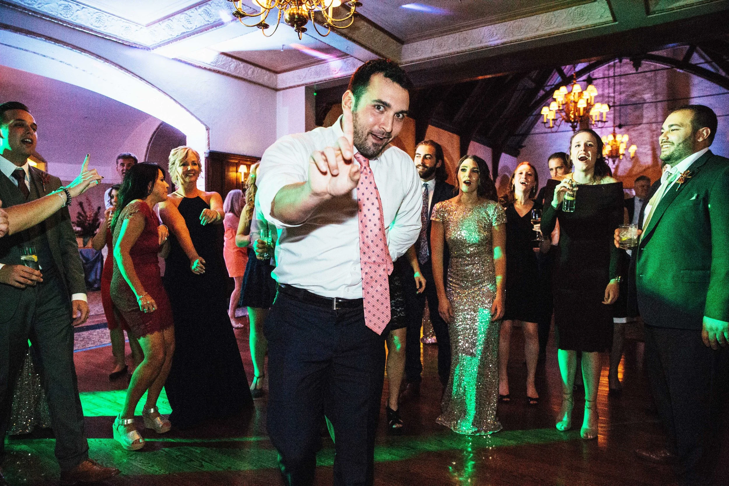 Wedding Guest dressed in formal attire dancing at a party with a man in the foreground making a playful gesture.