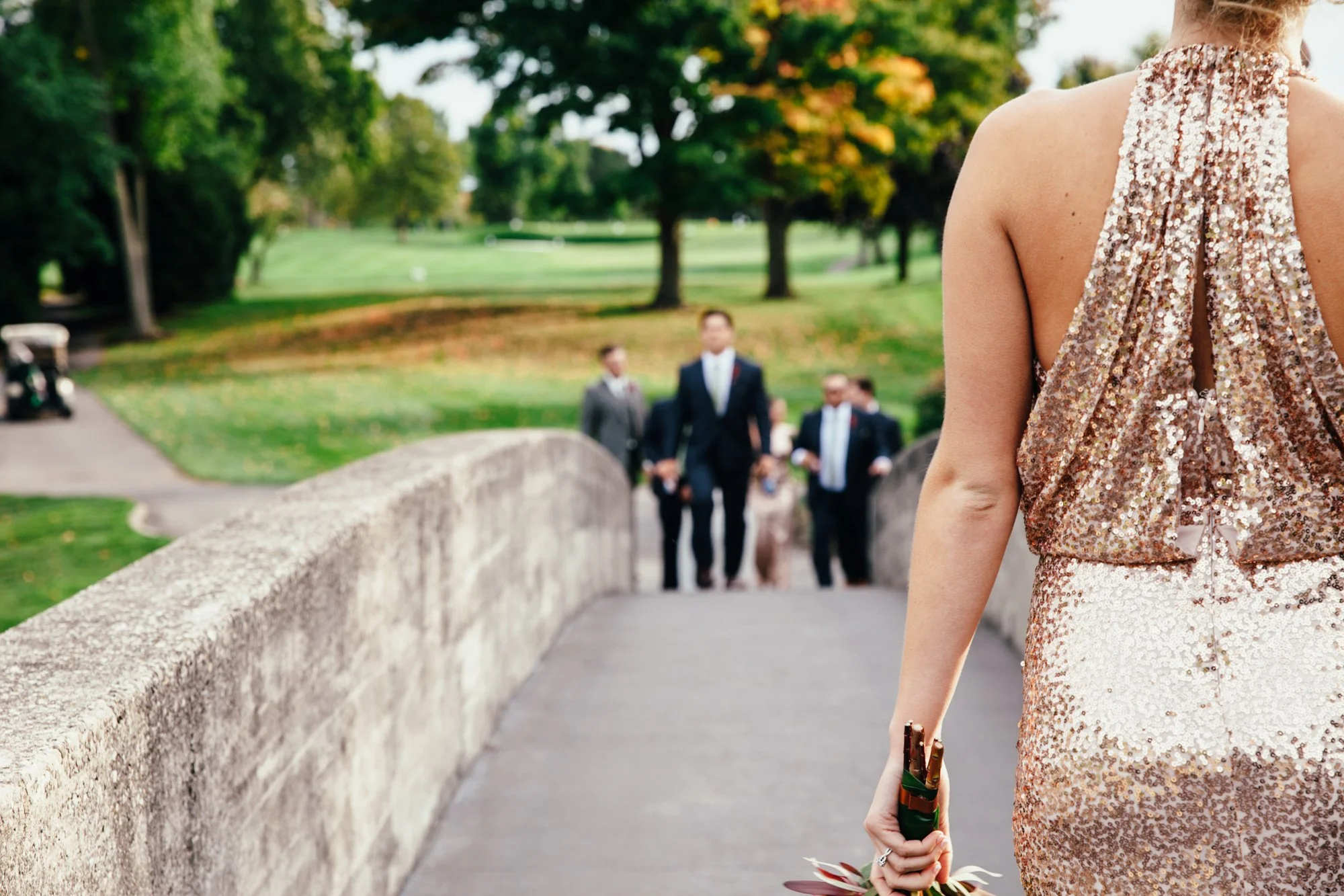 Bridesmaid in a sequined dress on a bridge holding a bouquet, with people in formal attire in the background and a golf course setting.