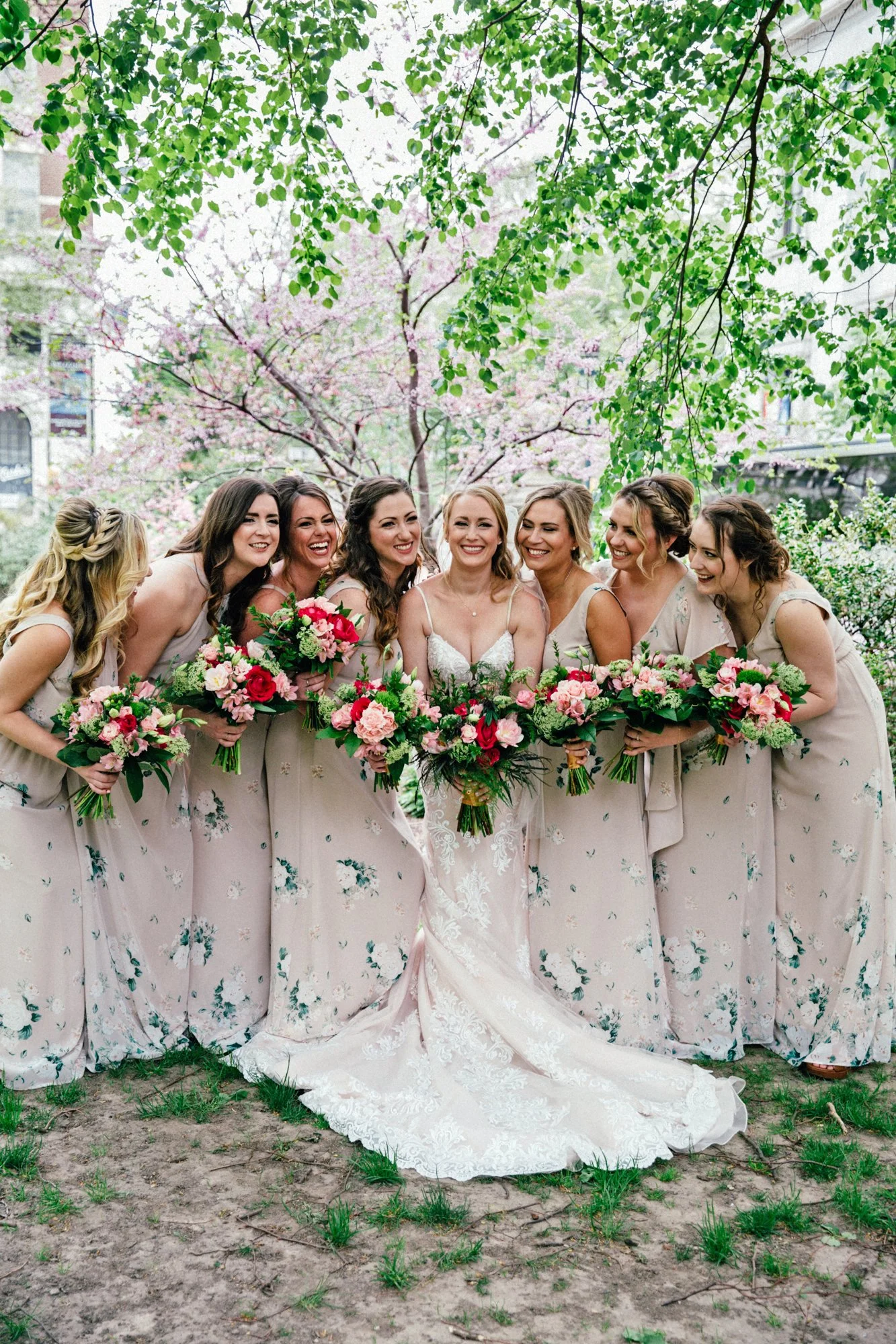 Bride with bridesmaids in floral dresses holding bouquets outdoors under trees.
