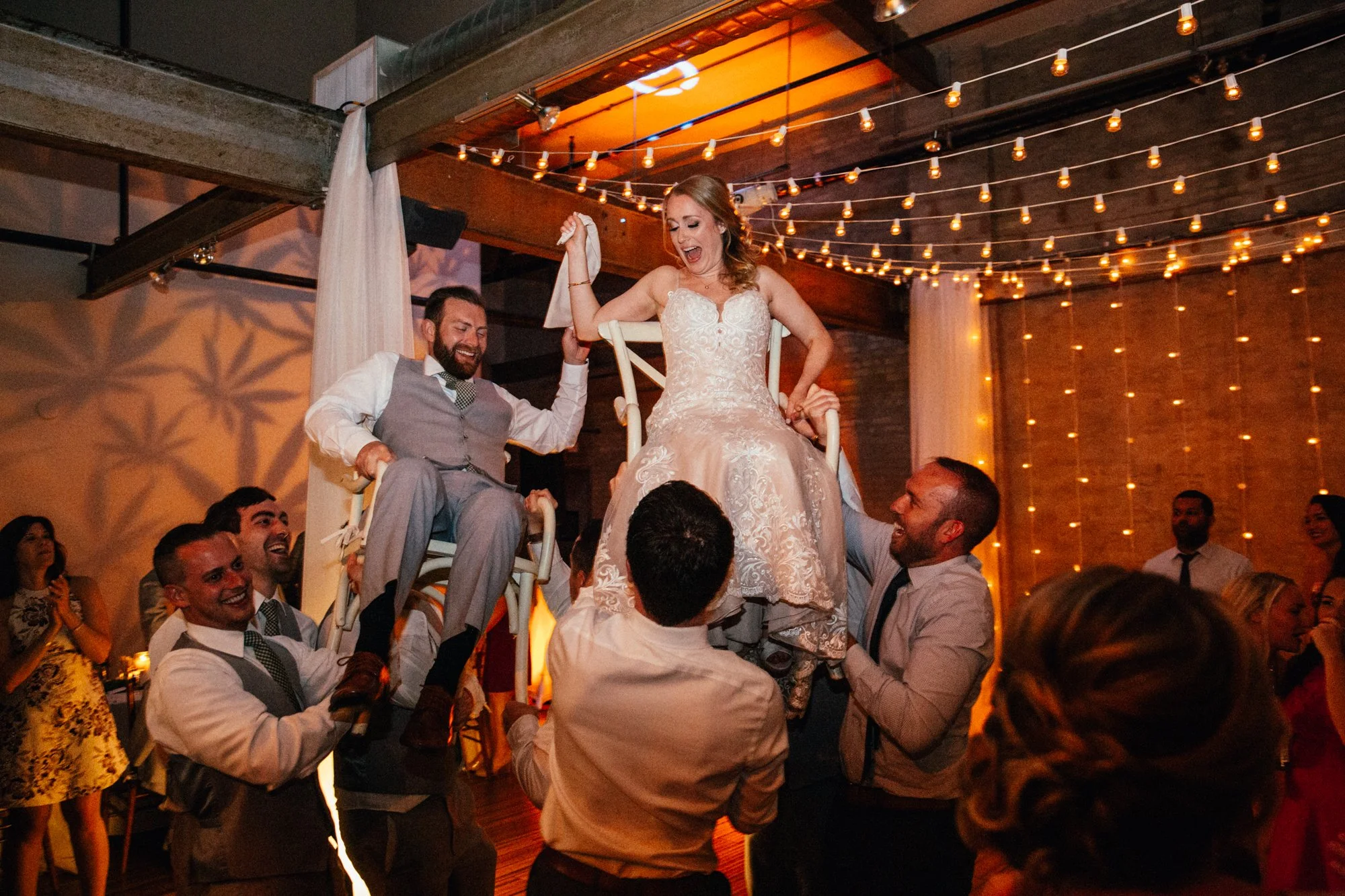 Bride and groom lifted on chairs during wedding celebration with string lights overhead.
