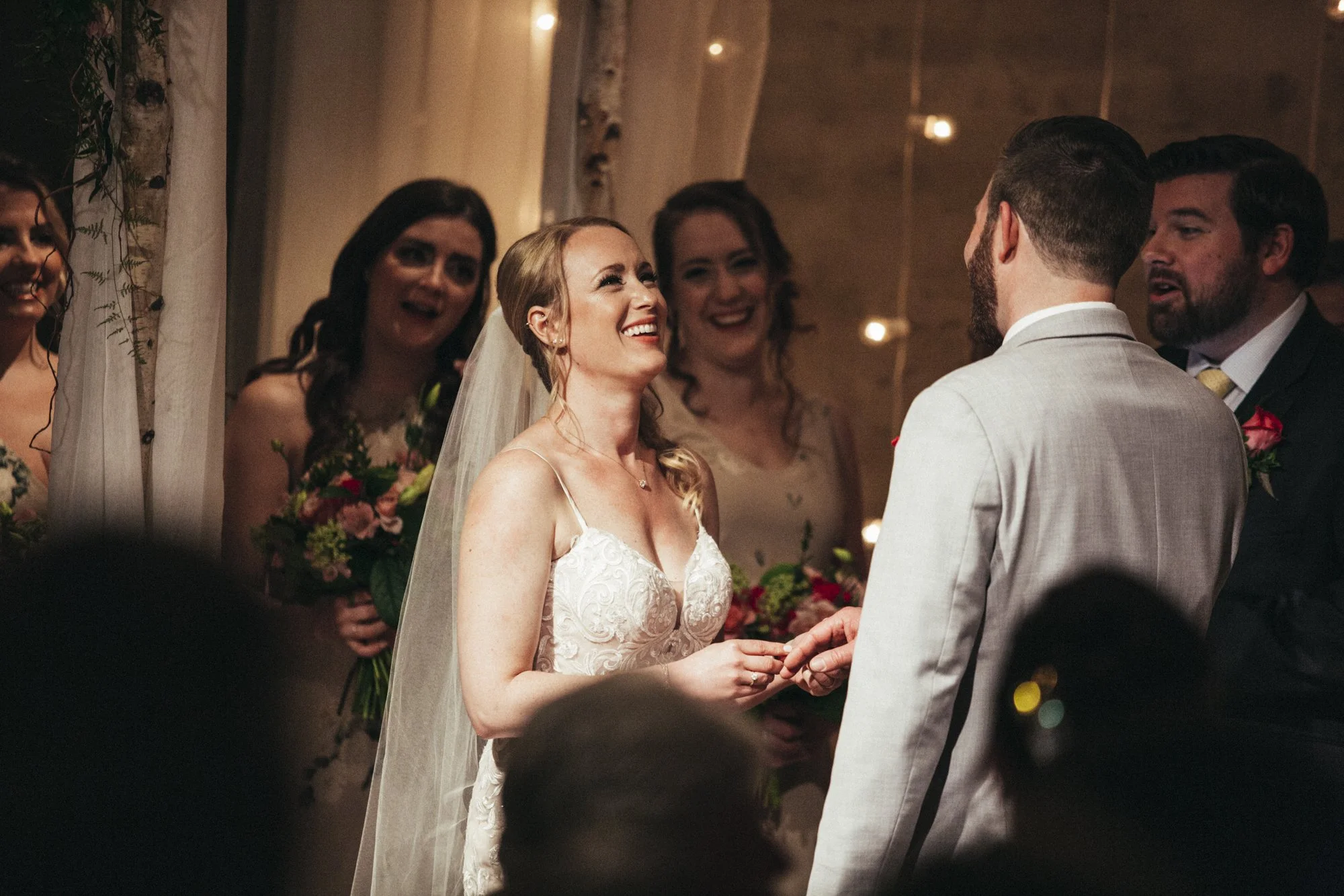 Bride and groom exchanging rings during wedding ceremony with bridesmaids and officiant in the background, decorated with flowers and string lights.