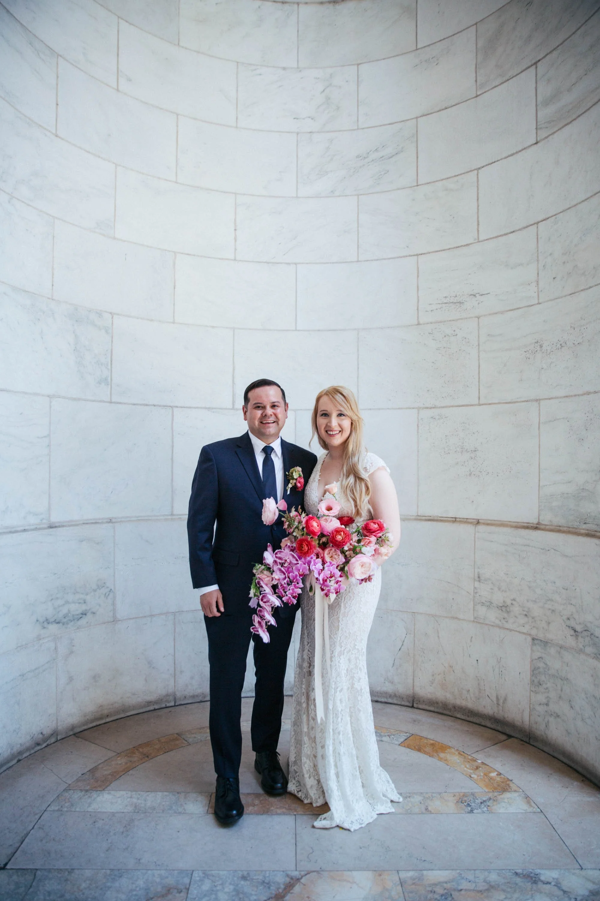 Bride and groom standing together, the bride holding a colorful bouquet, in a marble-walled setting.