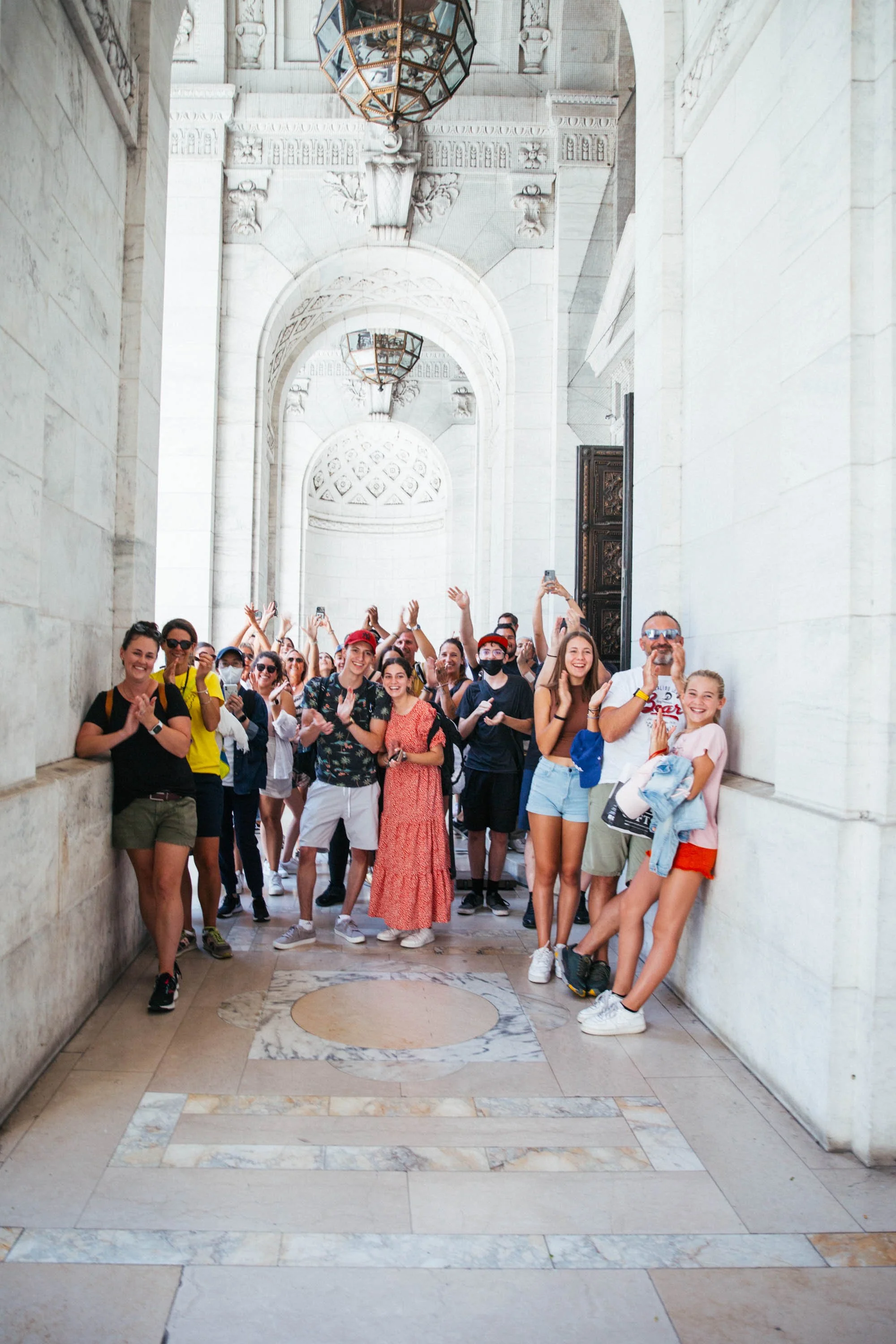 A group of tourists standing under the arches of the Public Library in New York City, clapping and smiling after discovering an elopement taking place.