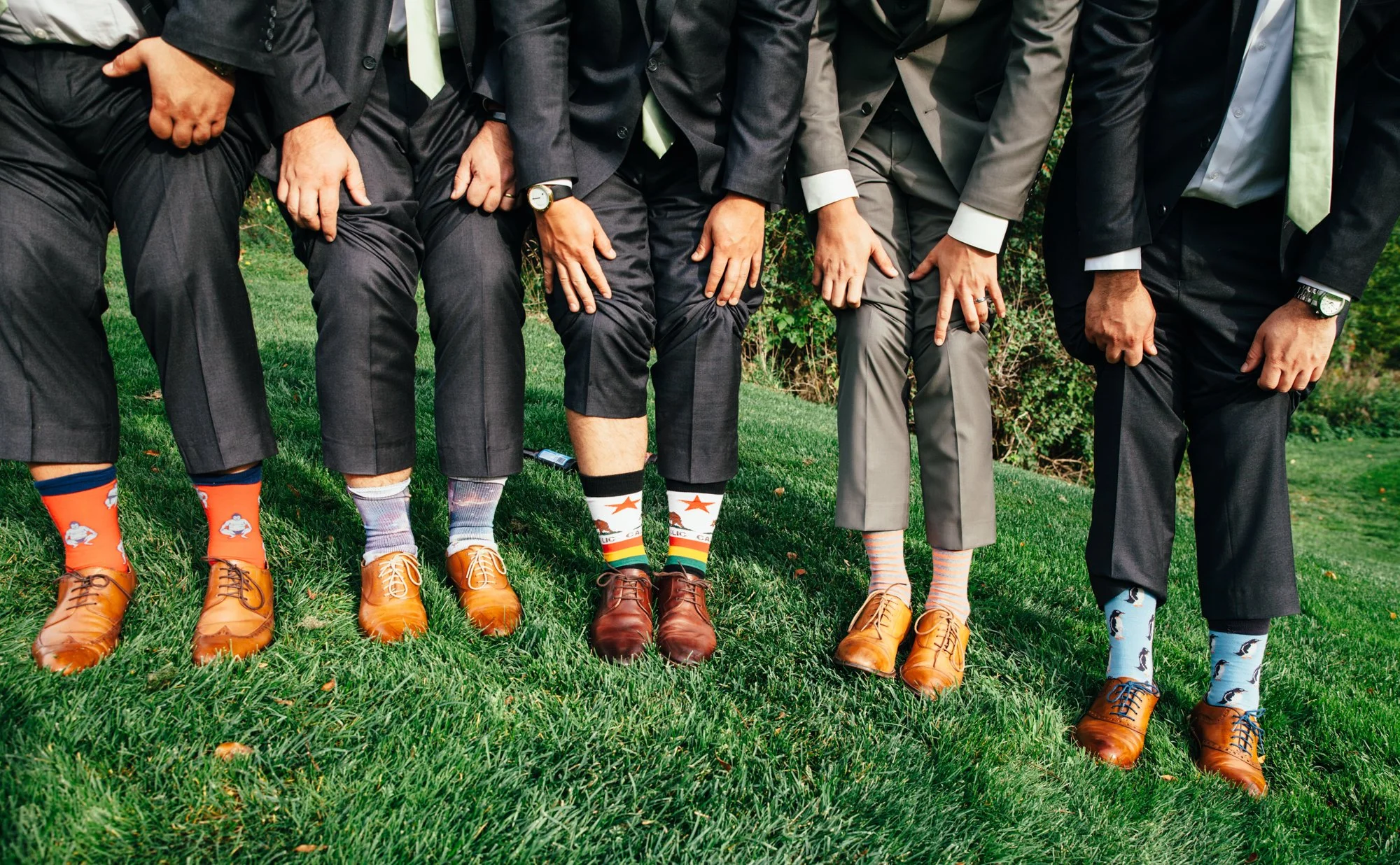 Groom and groomsmen in suits showing off colorful, patterned socks on grass.
