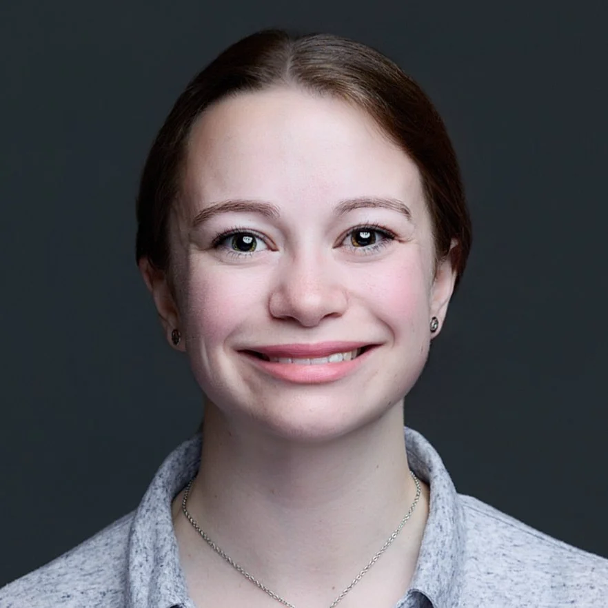 Portrait of Kelly Crawford with brown hair tied at the back of their head and light skin, smiling, wearing small round silver earrings, a silver necklace, and a light blue top against a dark background.