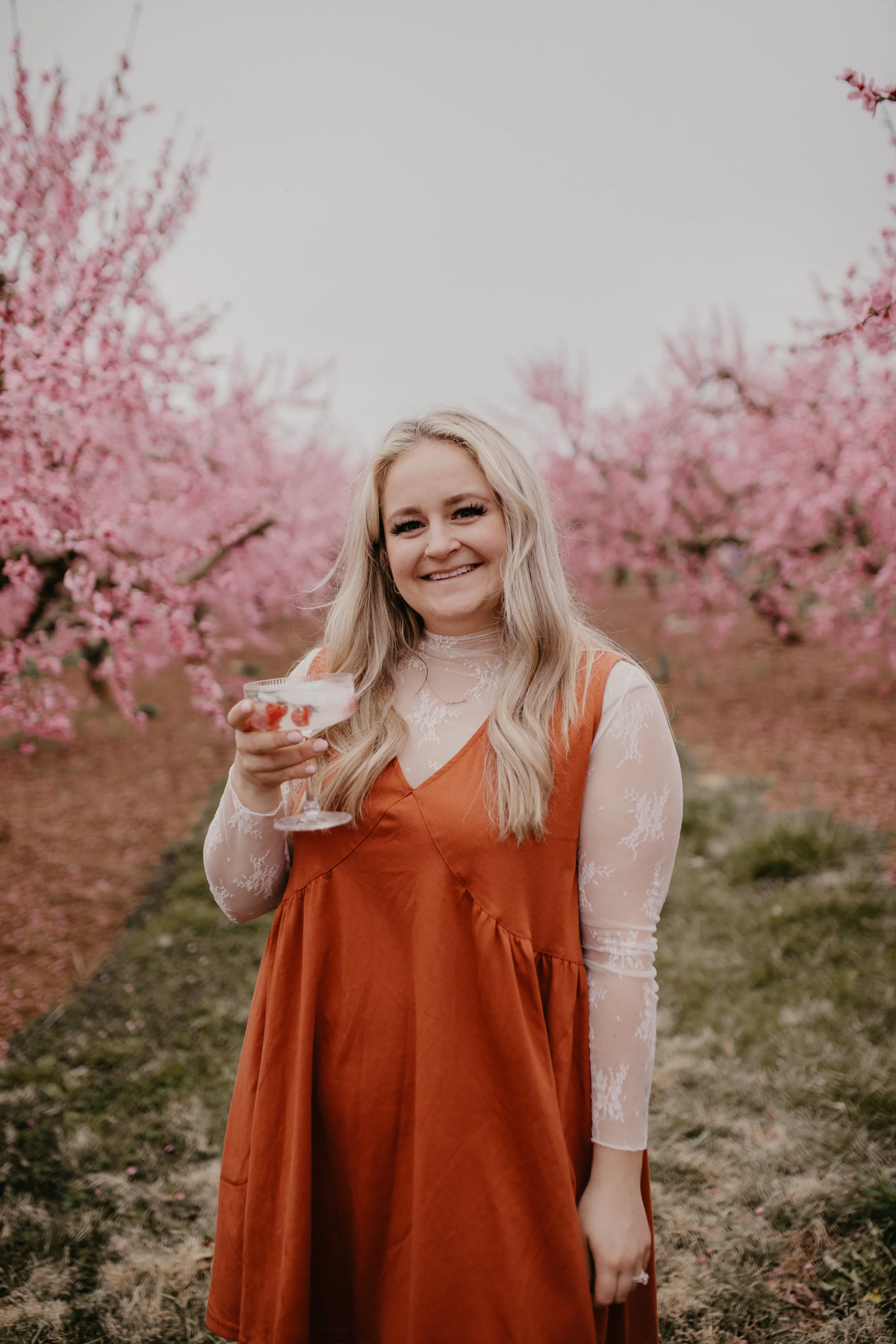 Woman in orange dress holding a drink in a pink flower orchard.