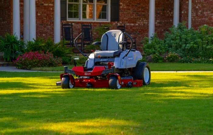 A red and gray riding lawn mower on a well-manicured lawn in front of a brick house with columns and a porch. Shrubs and flowers are visible in the background.