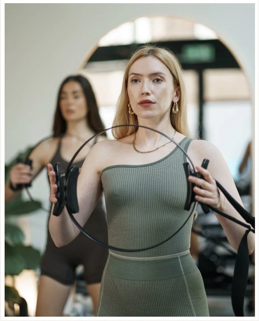 A woman with long blonde hair in a black dress using a rowing machine in a fitness studio, with a mirror reflecting her and others exercising.